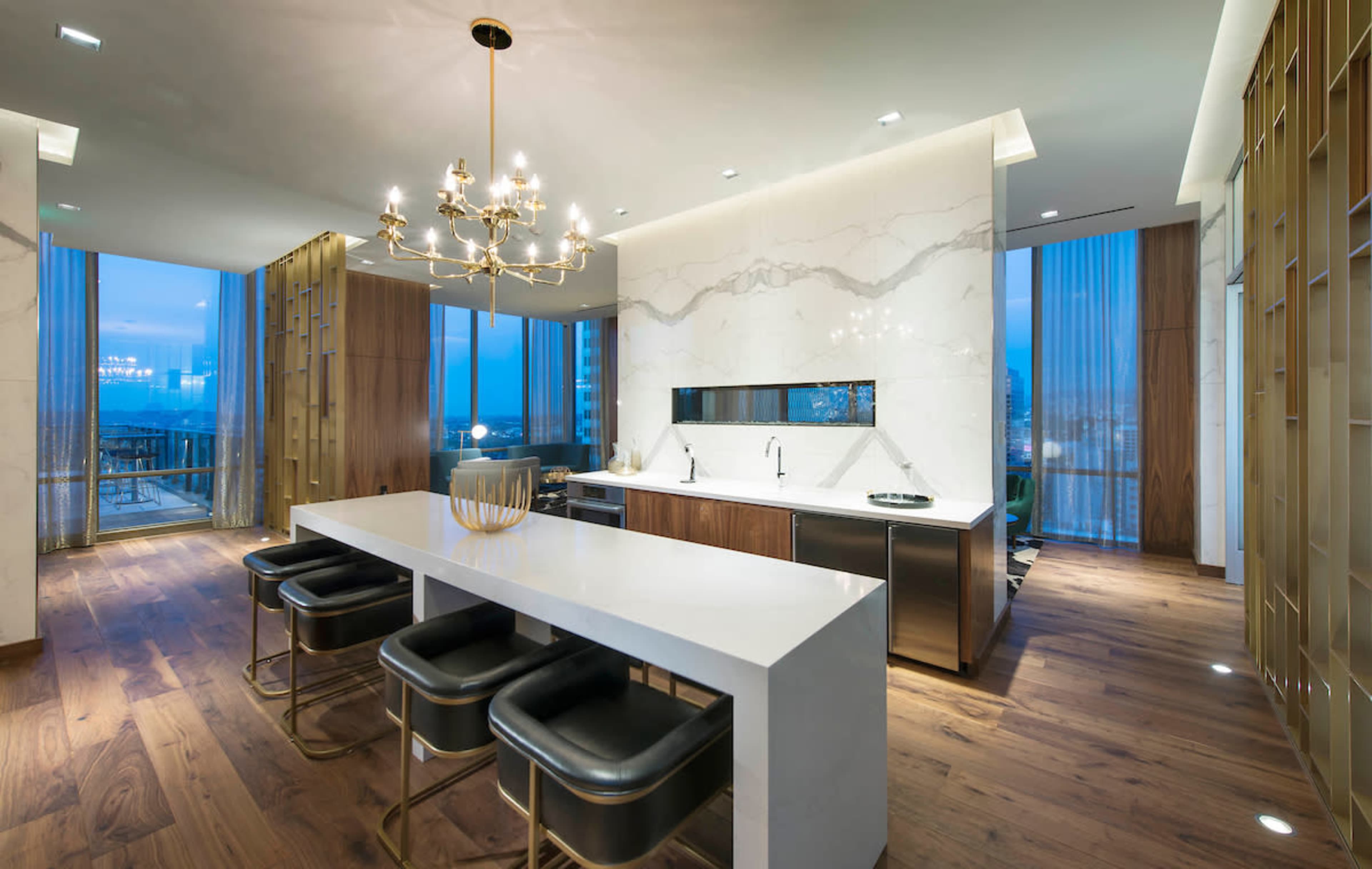 The image shows a modern kitchen with a large white island, sleek cabinetry, and a chandelier, illuminated by natural light from floor-to-ceiling windows.