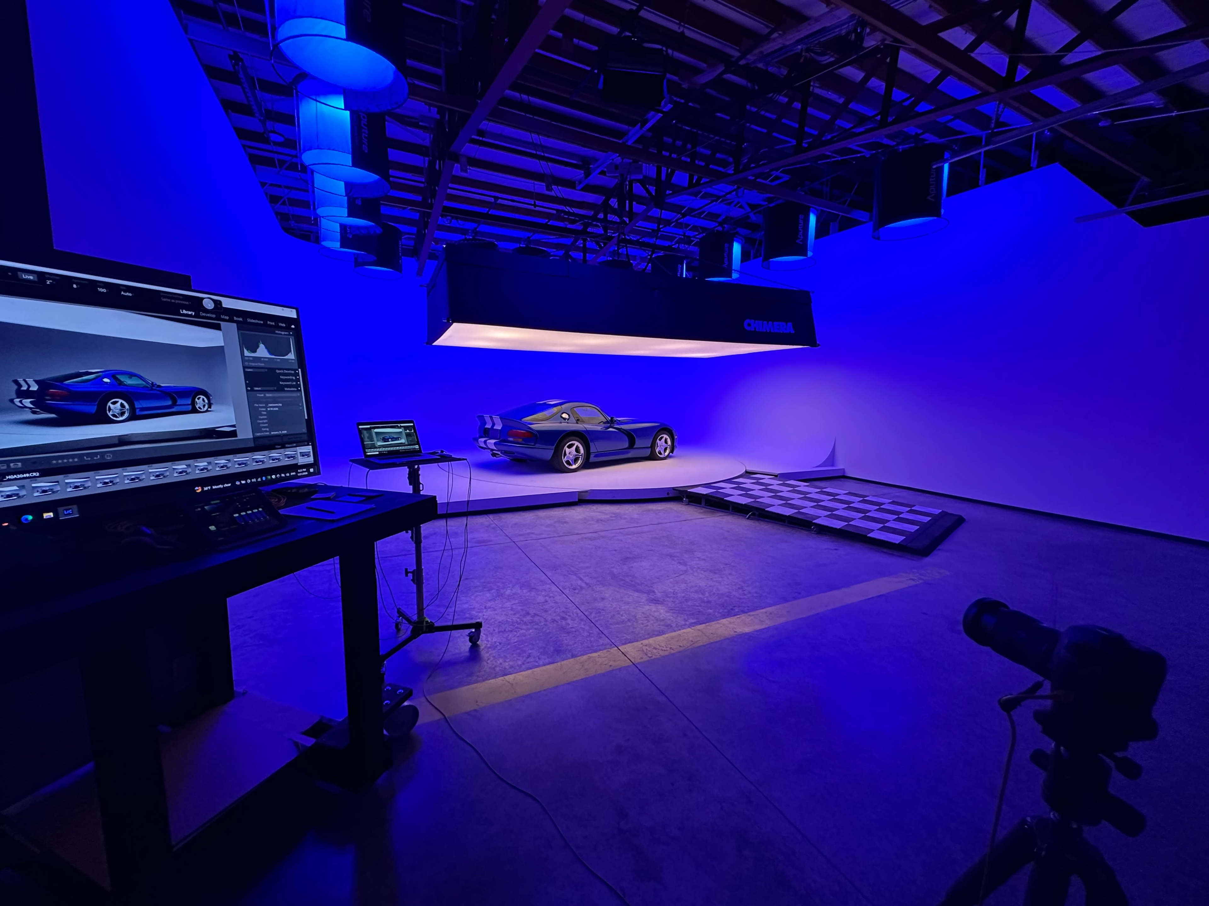 A blue-lit photography studio showcases a car on a display platform, with computer equipment visible in the foreground.