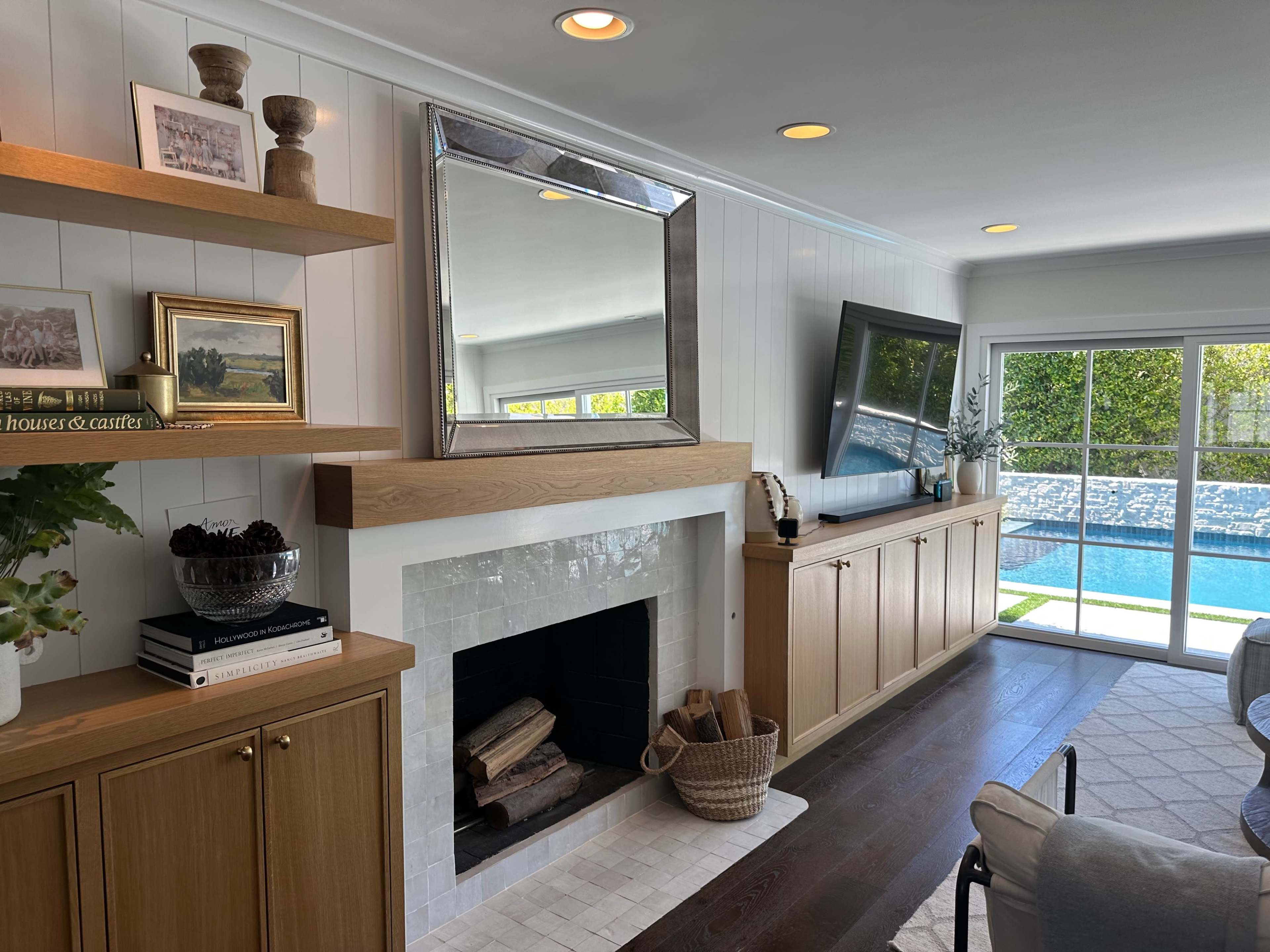A modern living room with a fireplace, a large mirror above it, built-in cabinetry, and a view through sliding glass doors to a swimming pool area.