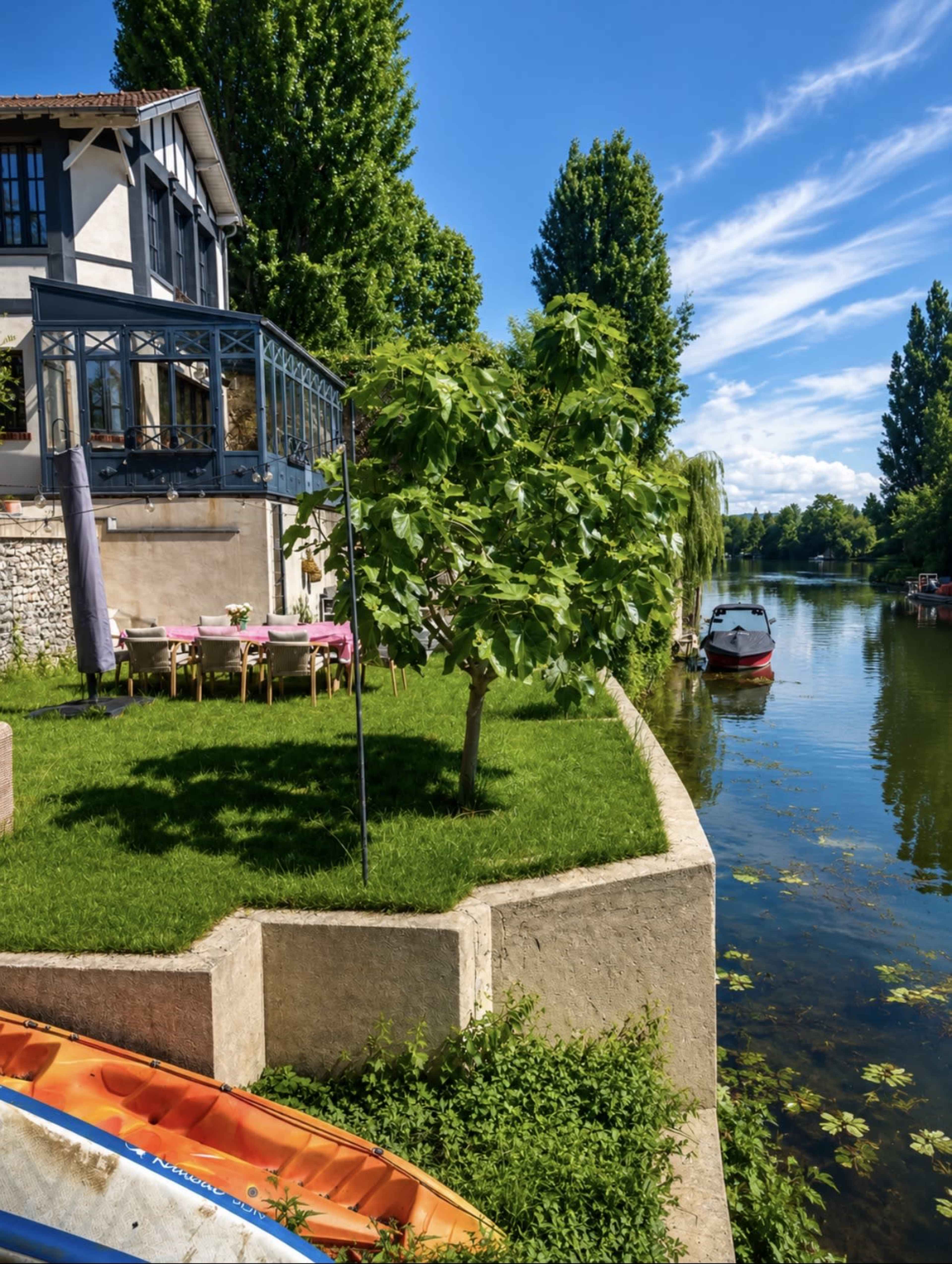 A well-maintained riverside property features a green lawn, a wooden deck, and a small boat docked along the water.