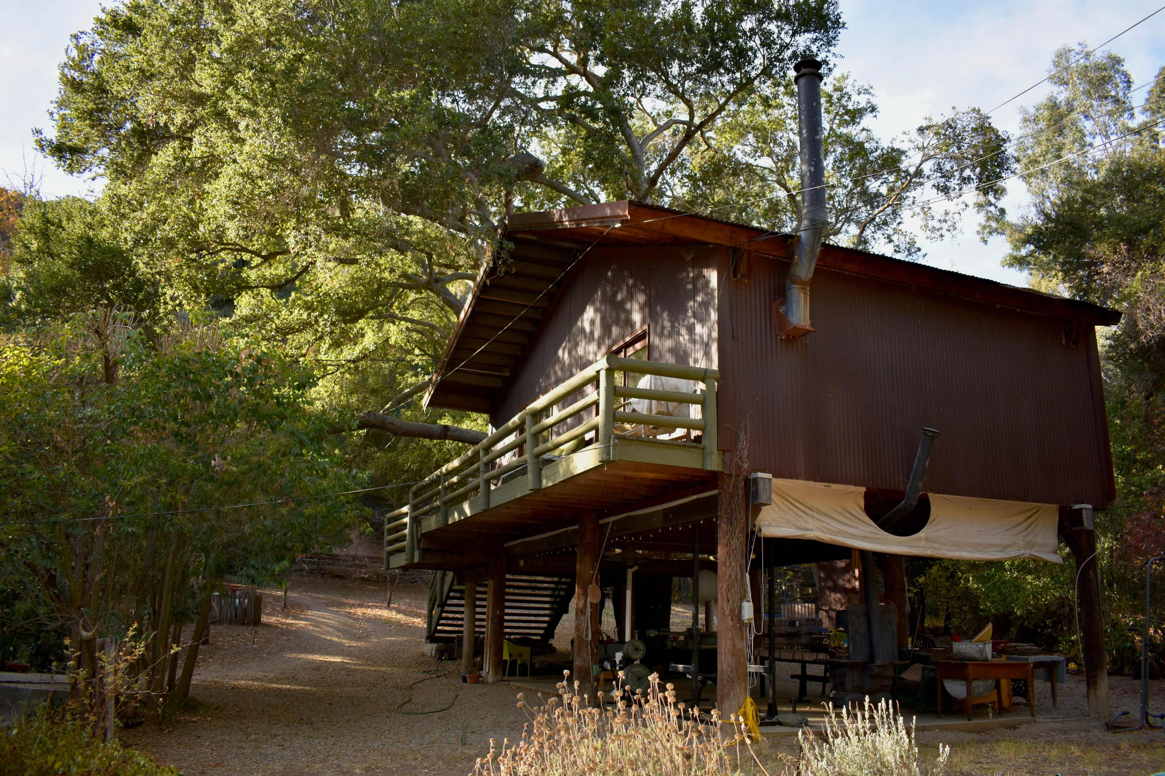A wooden house elevated on stilts sits under large trees, with a spacious deck and outdoor seating area below.