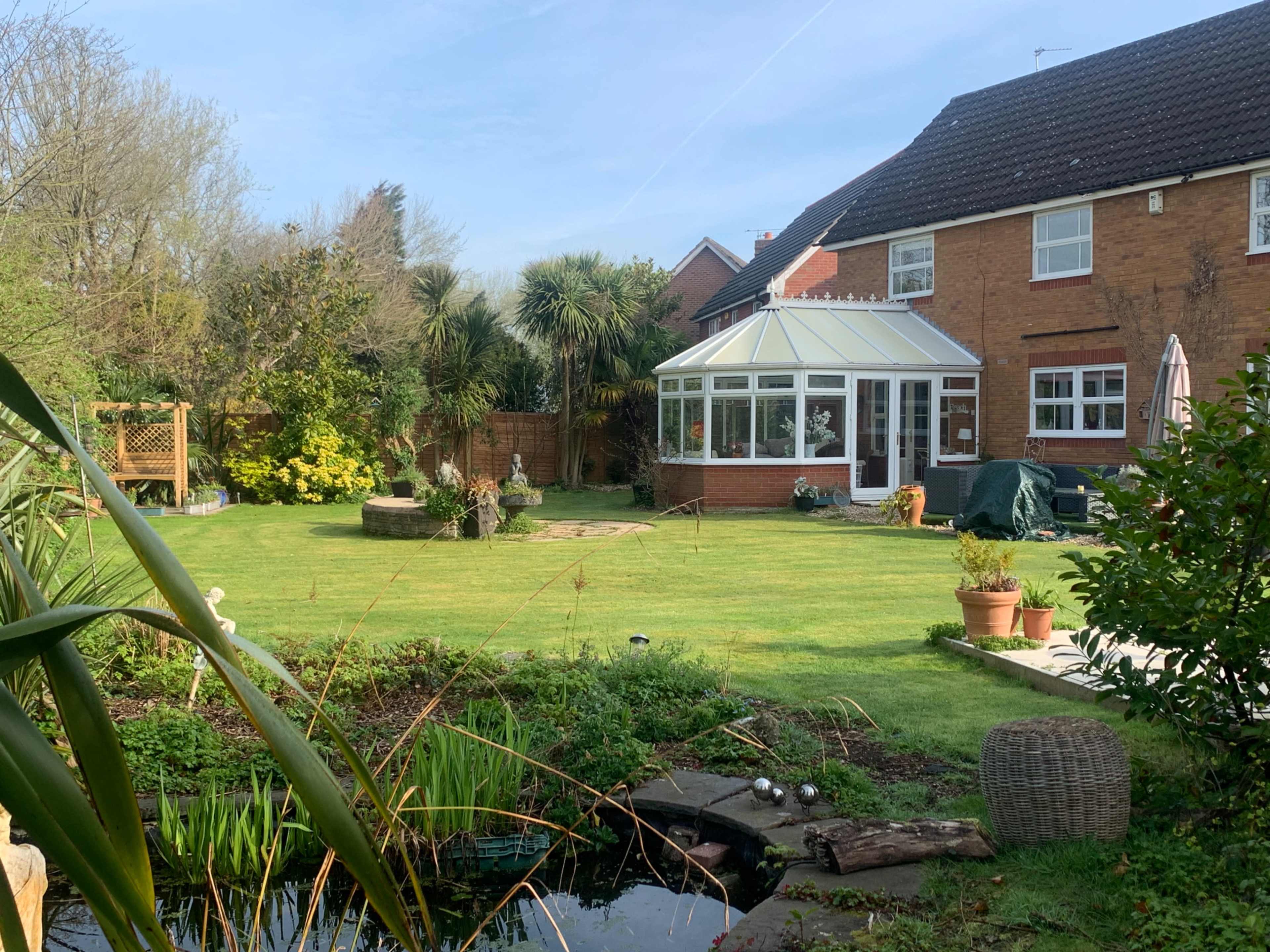 The image shows a well-maintained backyard featuring a grassy area, a conservatory, various plants, and a small pond.