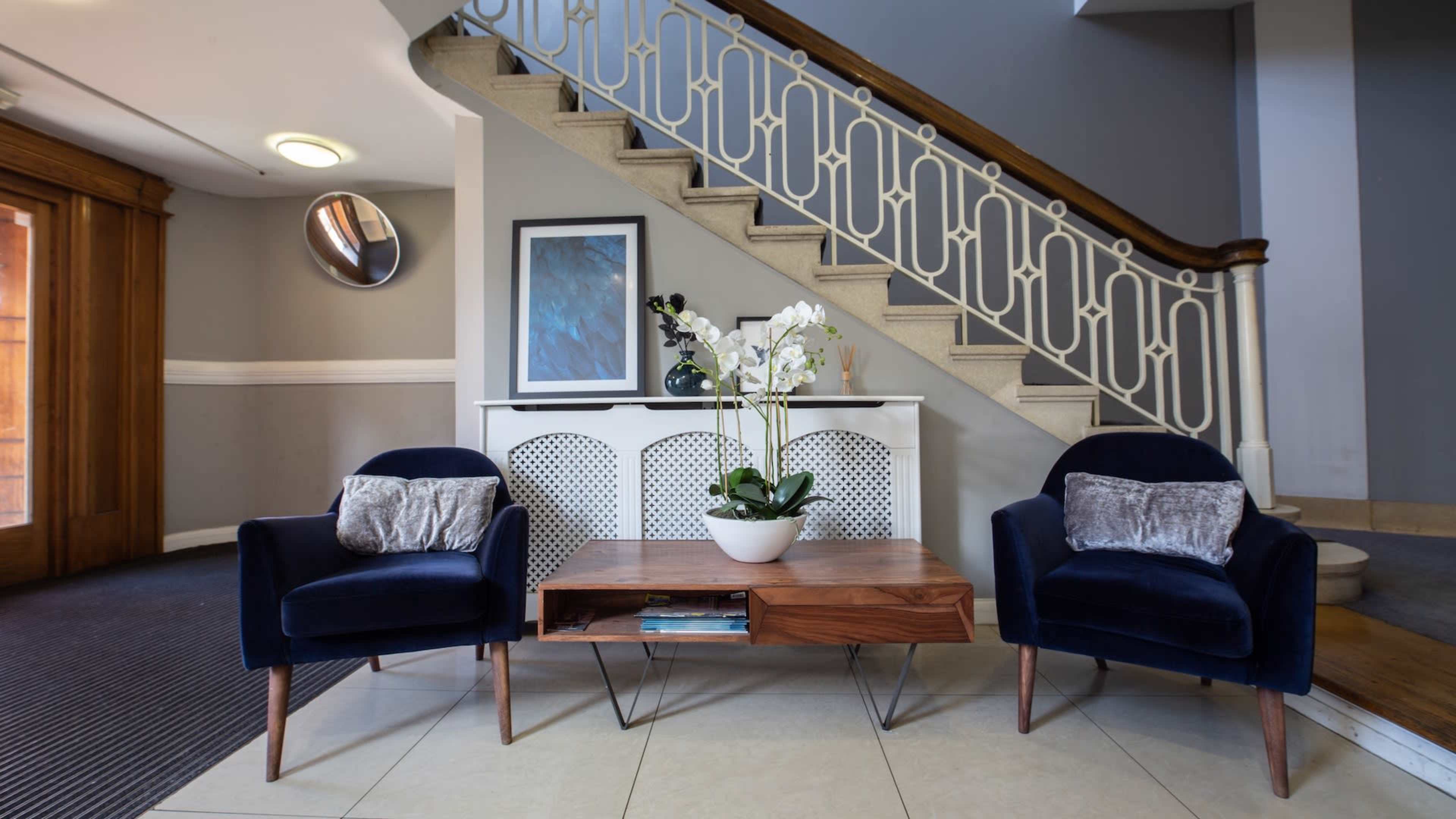 A modern lobby features a wooden side table with a potted orchid, flanked by two navy blue chairs, beneath a staircase with decorative railings.