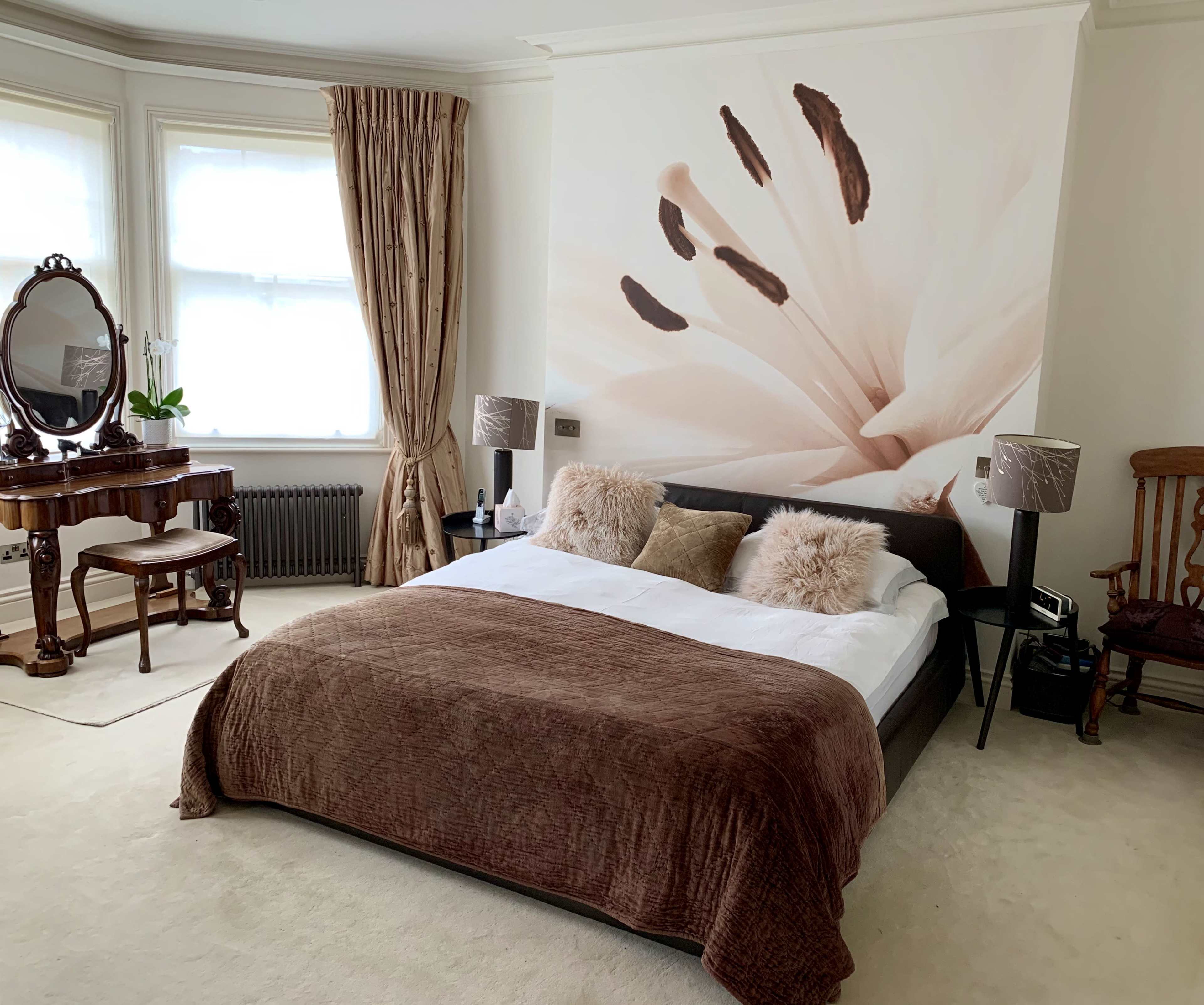 A spacious bedroom featuring a large bed with a brown blanket and decorative pillows, flanked by two bedside lamps, a vintage vanity table, and a floral mural on the wall.