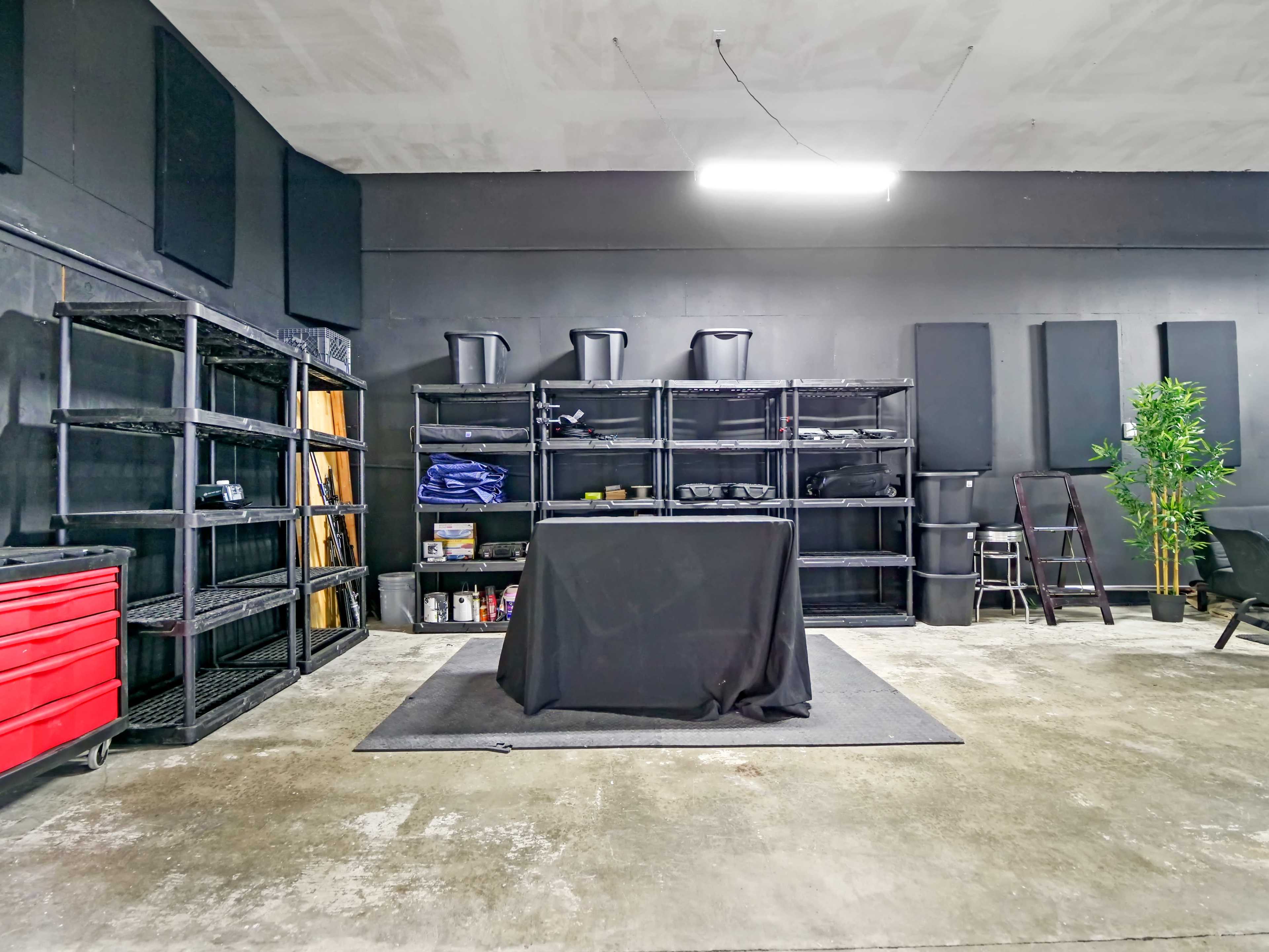 A spacious room with black walls, shelving units, a central table covered with a black cloth, and storage containers, along with a potted plant in one corner.