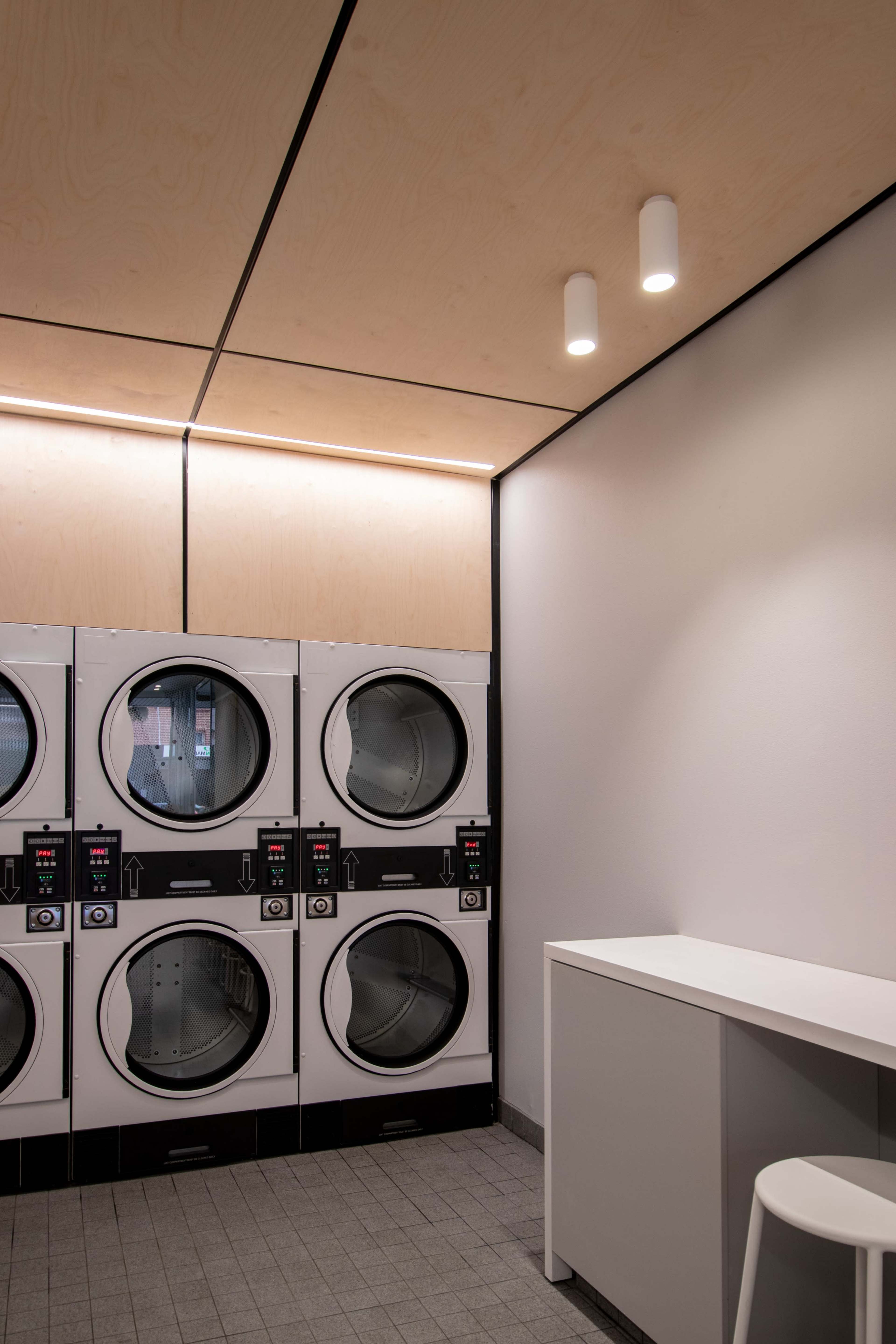 A laundry room features stacked washing machines and dryers against a light-colored wall with minimalist lighting.