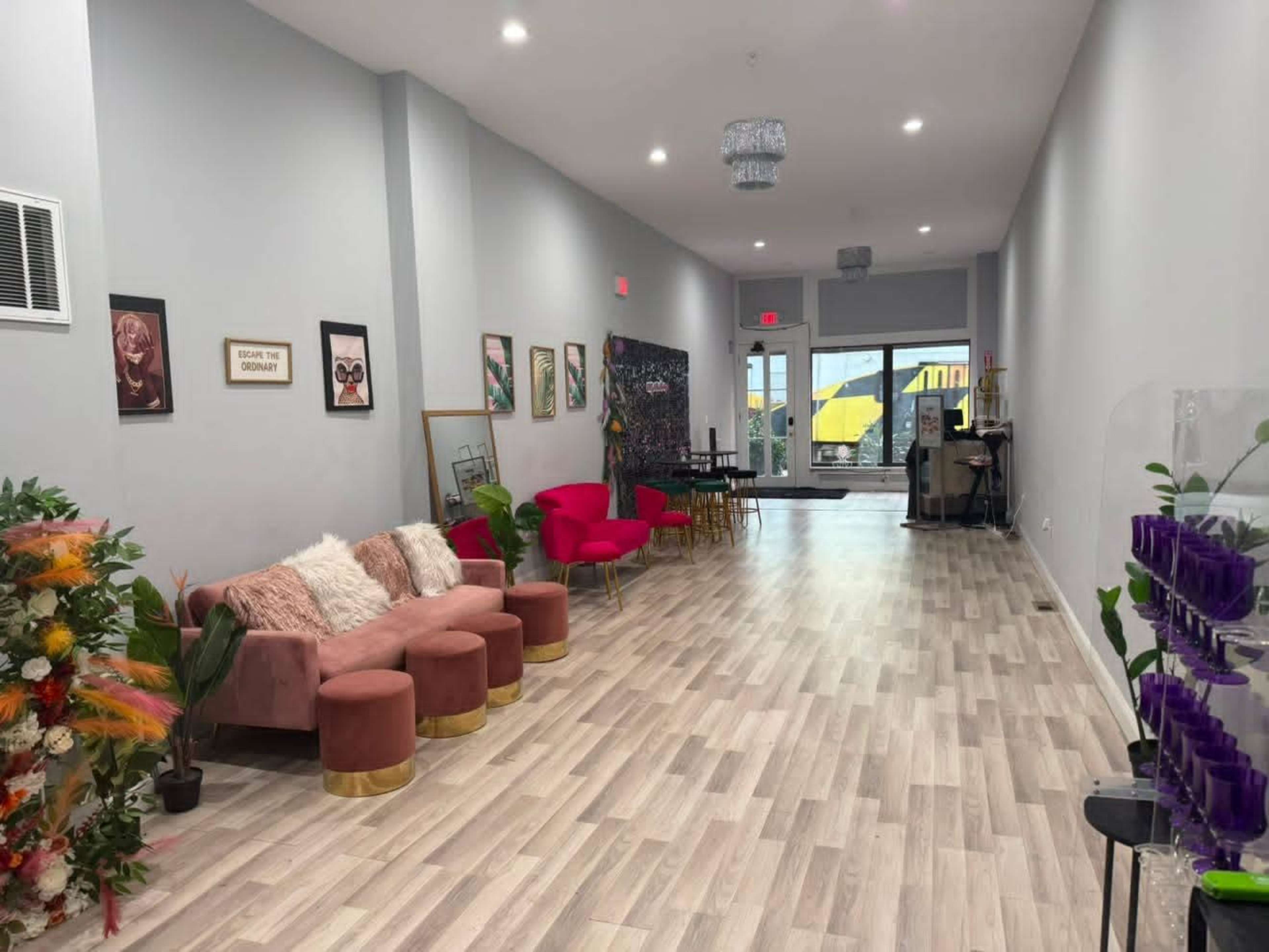 The image shows a modern, spacious interior with pink seating, artwork on the walls, and a bright entrance leading to the outside.