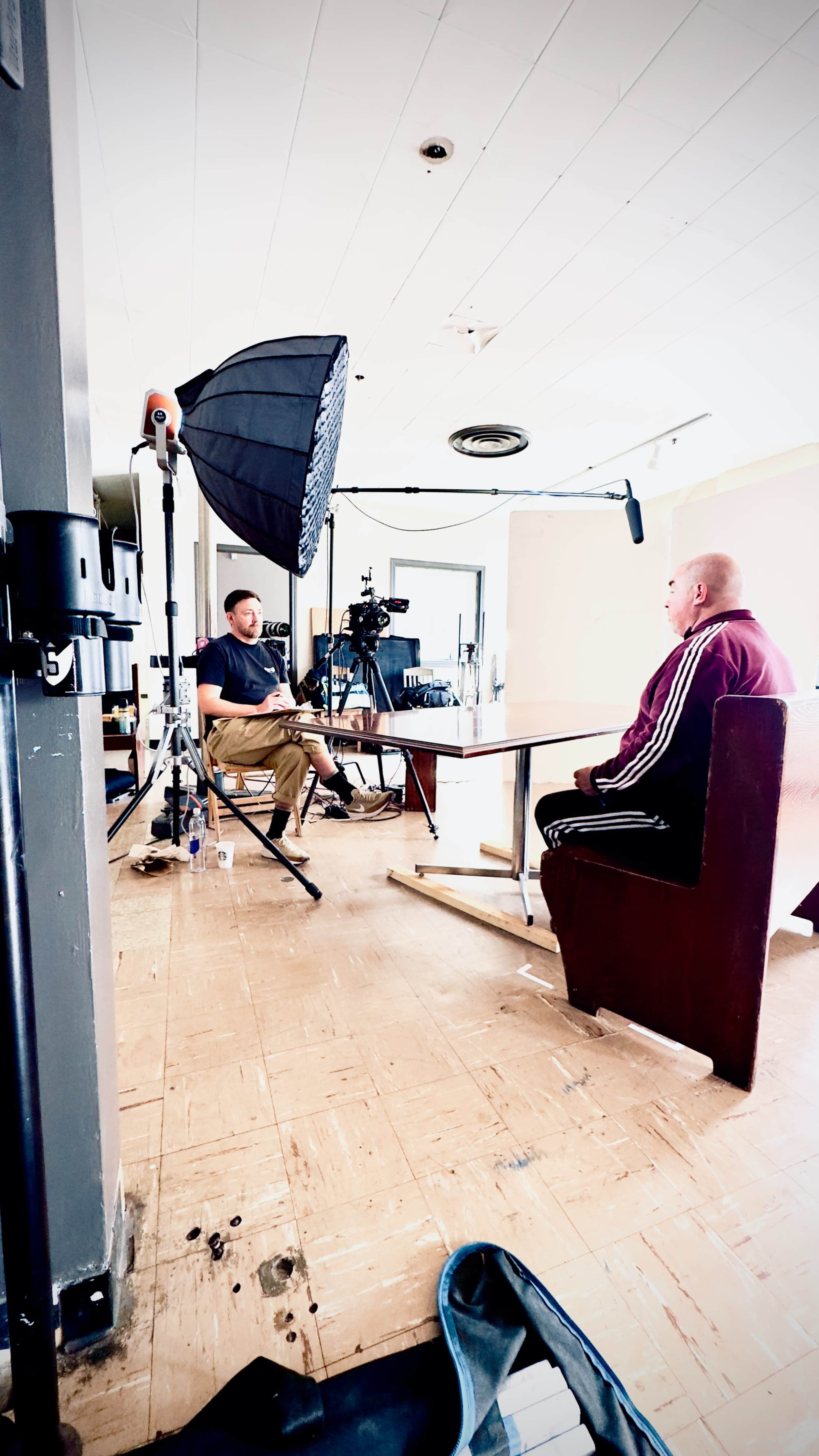 A man sits at a table facing another man, who is filming the interview with a camera and lighting setup in a well-lit room.