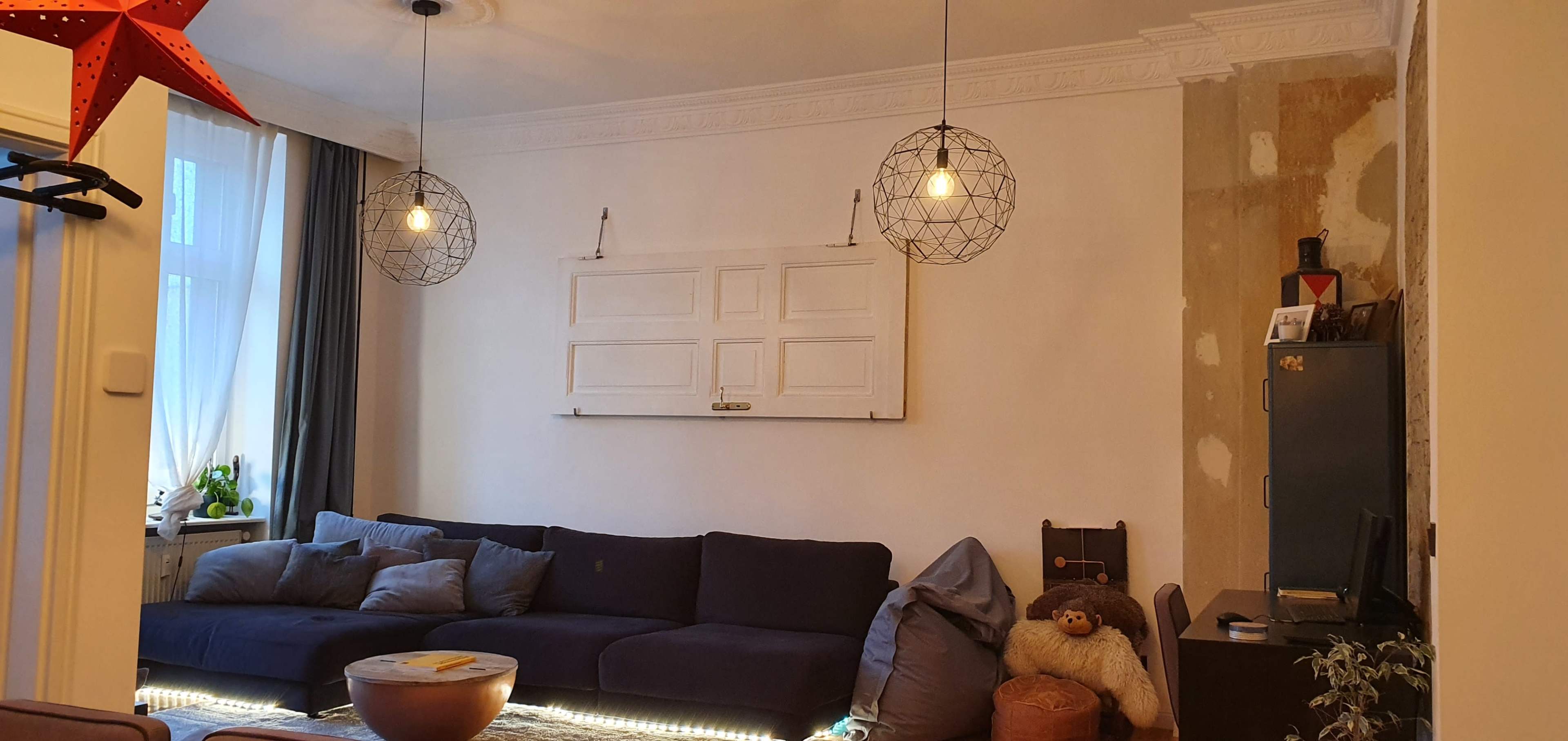 The image shows a living room with a dark sofa, two pendant lights, a decorative wall with an old door frame, and various furniture pieces.