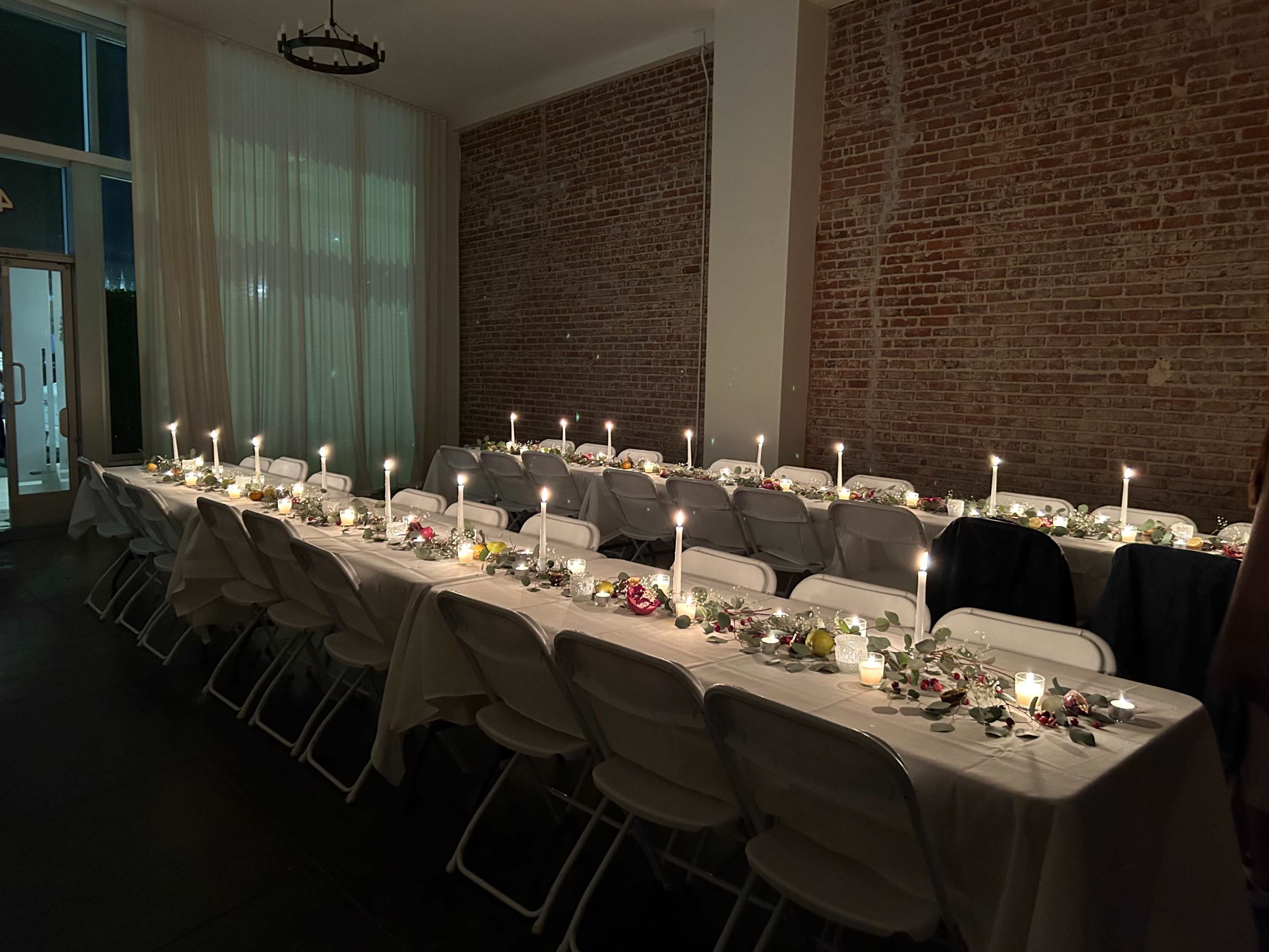 A dimly lit dining room features long tables adorned with floral decorations and lit candles.