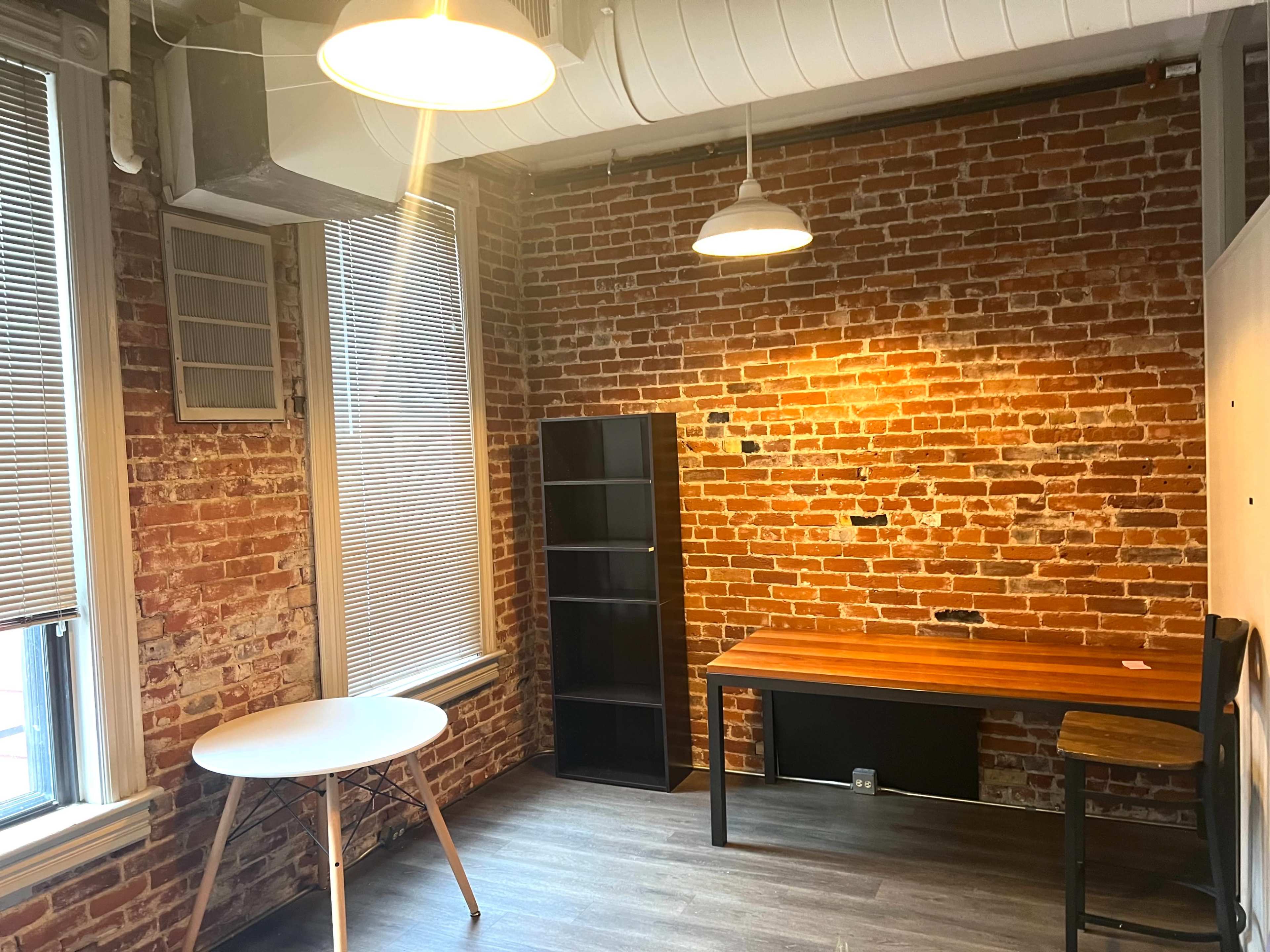 A room with exposed brick walls, a round white table, a wooden desk, and a black bookshelf.