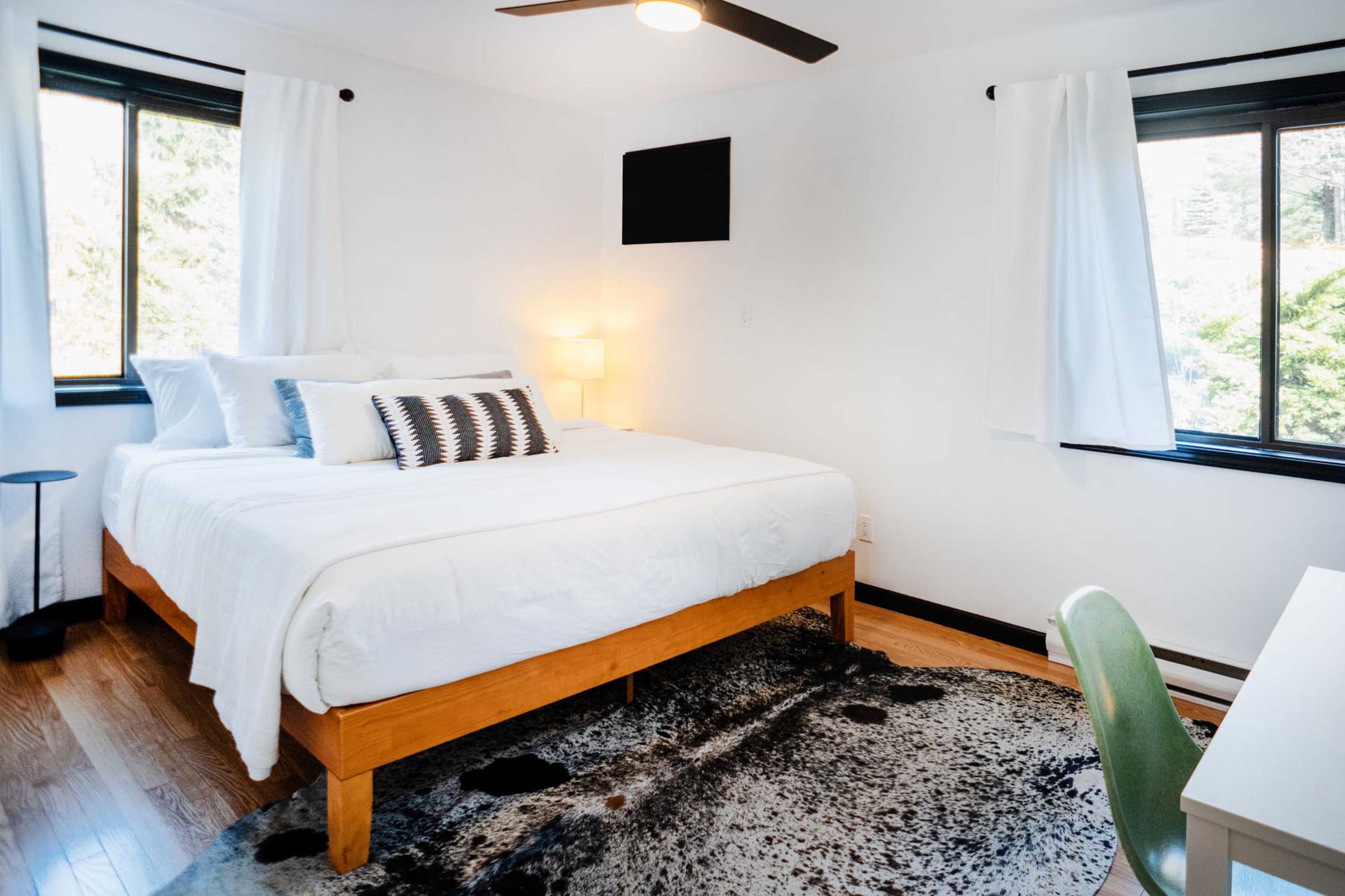 A neatly arranged bedroom with a large bed, a small bedside lamp, and a desk chair placed on a patterned rug.