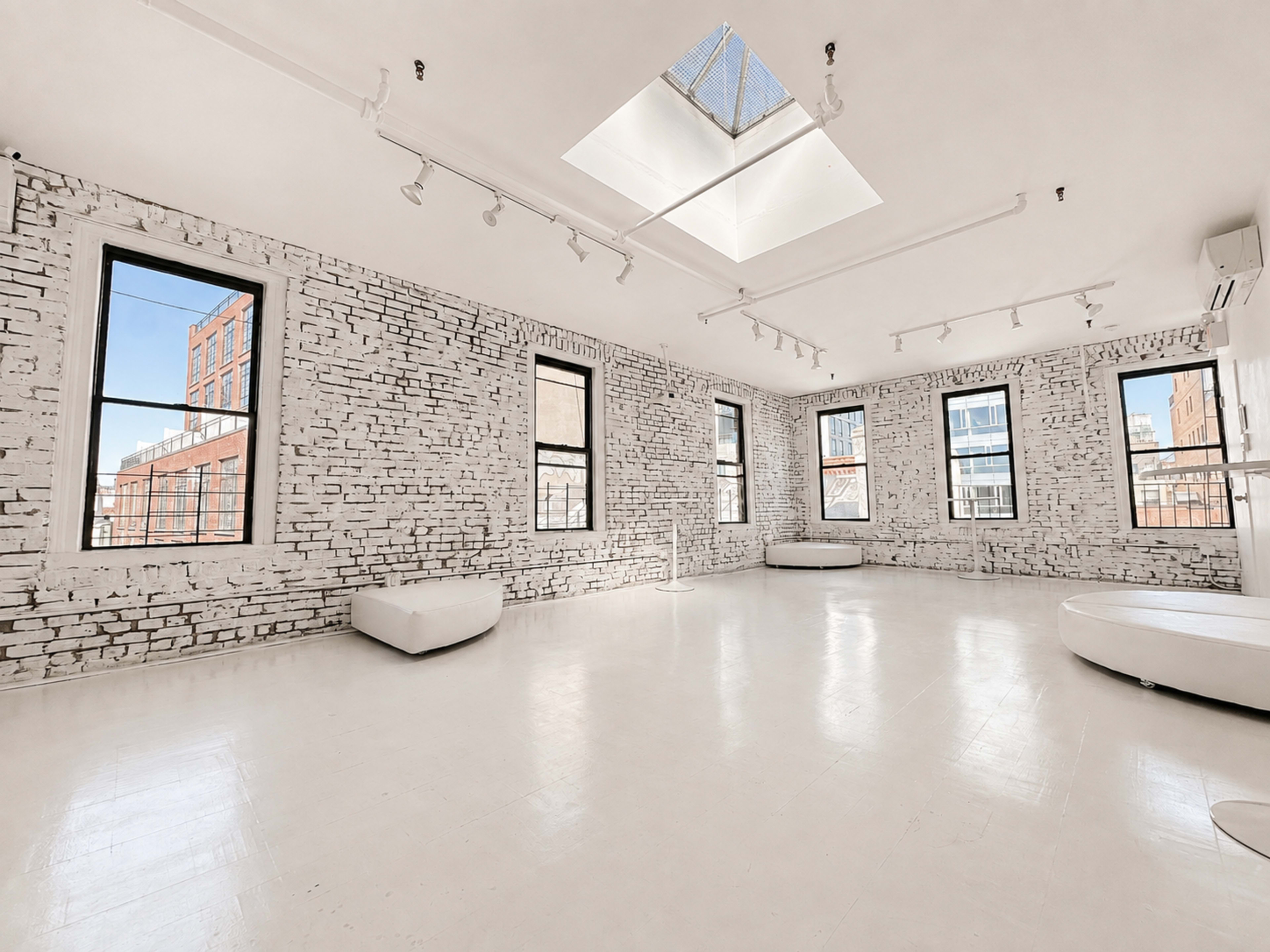 The image shows a bright, minimalistic studio space with white brick walls, large windows, and a skylight, featuring a polished white floor and two circular seating areas.