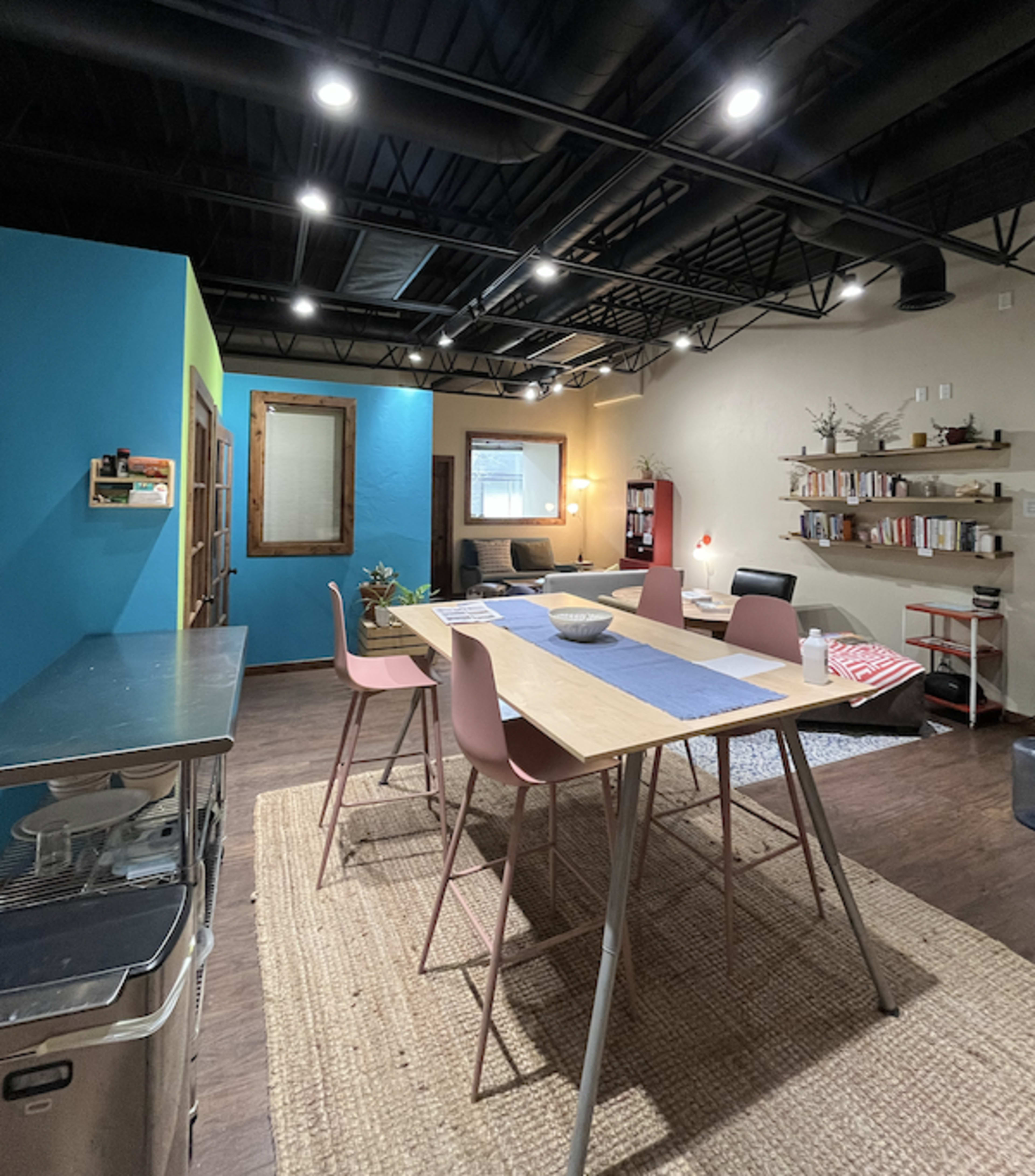 The image shows a modern, open-plan kitchen and dining area with a wooden table, pink chairs, and shelves filled with books, set against brightly colored walls.