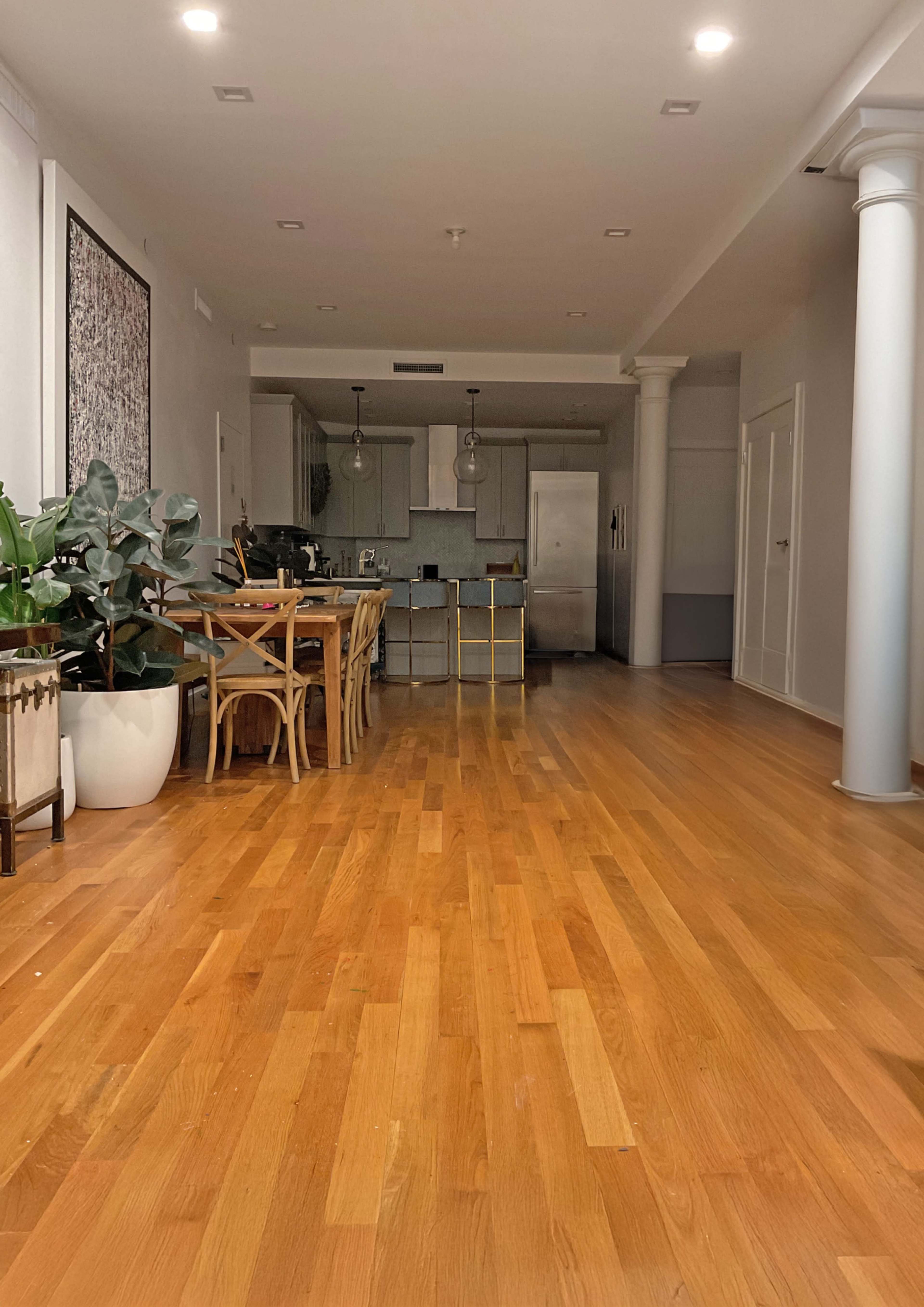 The image shows a spacious wooden-floored room with a kitchen in the background, featuring a dining table and plants along the walls.