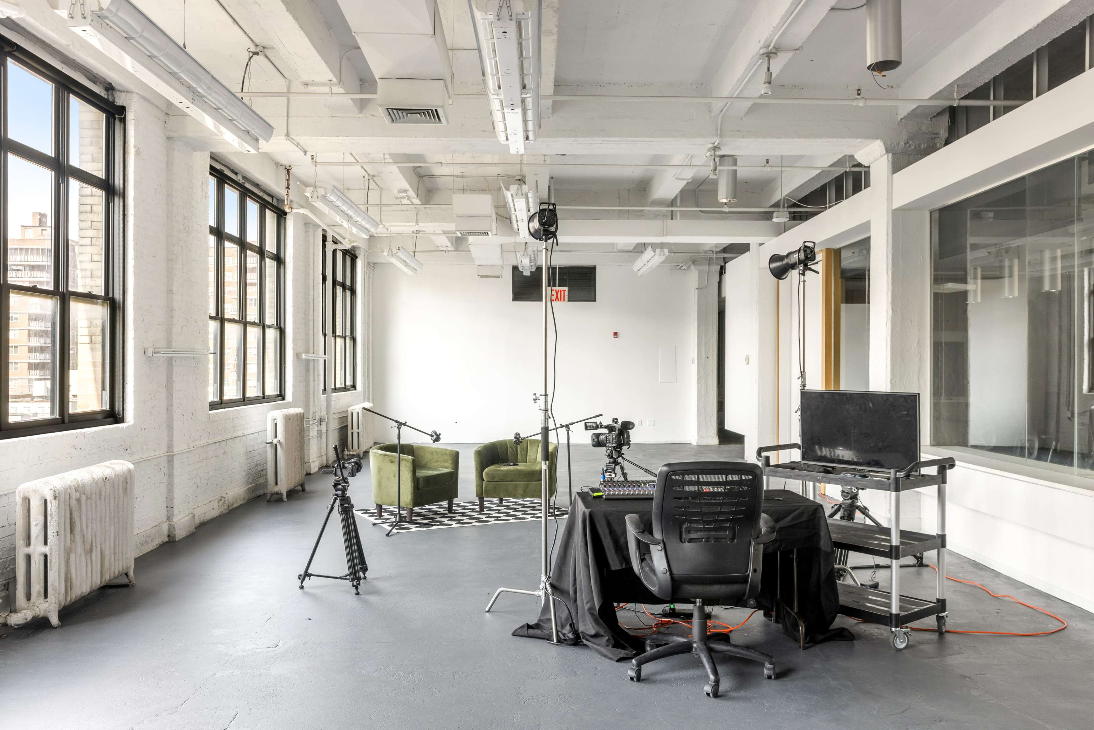 The image shows a spacious, modern office with large windows, a black and white patterned rug, green seating, and equipment set up for video recording.