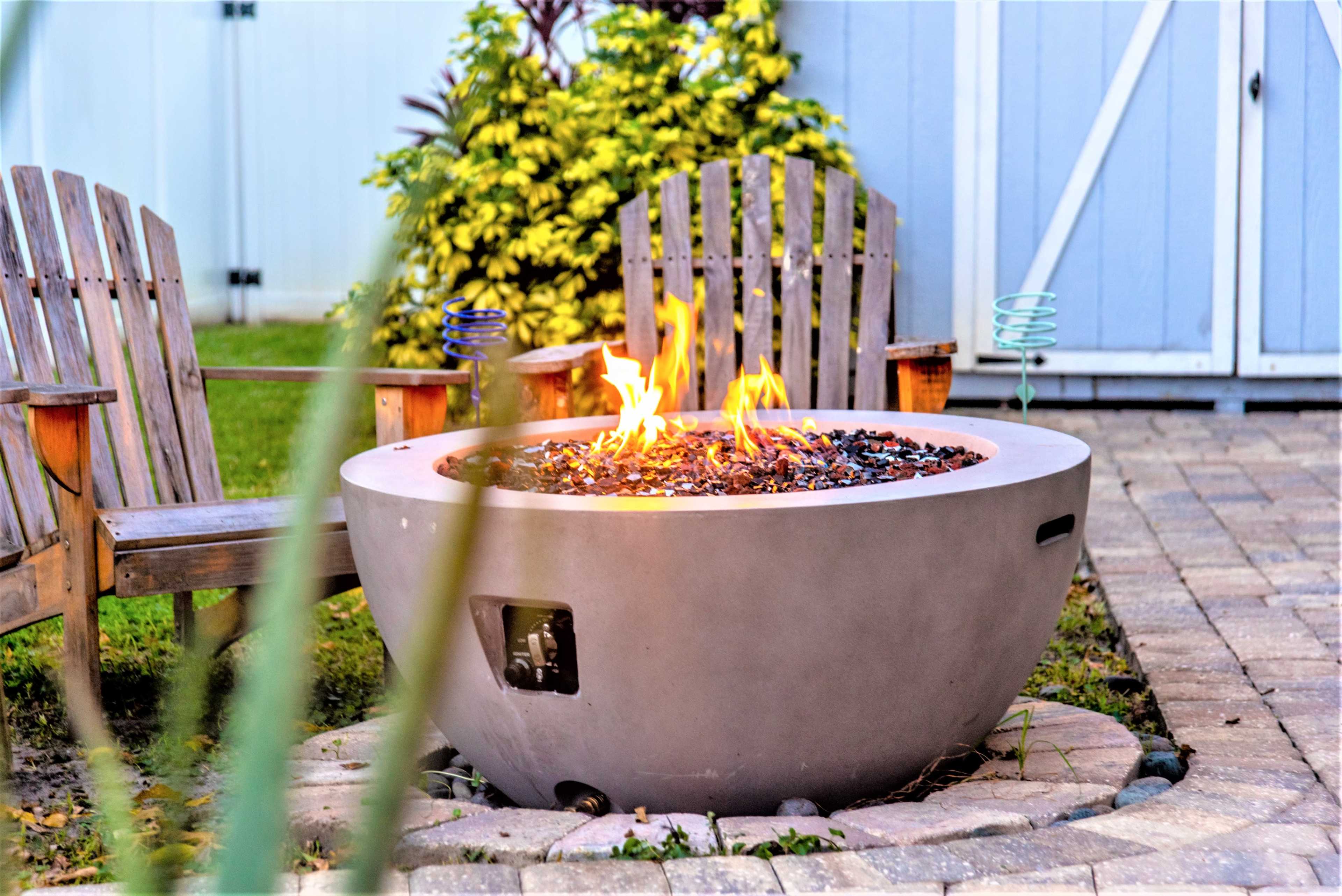 A round fire pit with flames is surrounded by two wooden Adirondack chairs in a landscaped backyard.