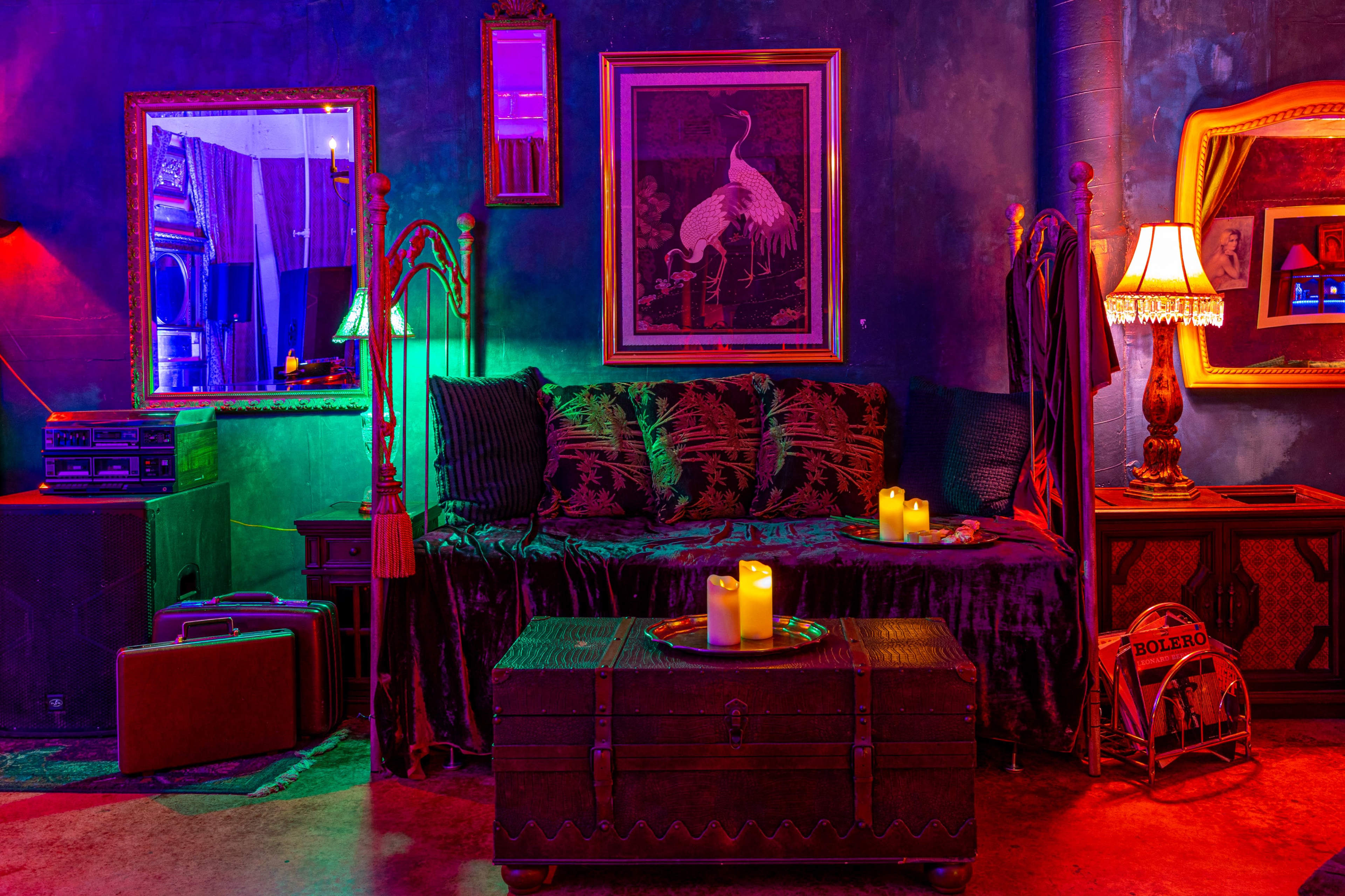 A dimly lit room featuring a plush sofa, decorative mirrors, a trunk, and illuminated candles on a table, surrounded by colorful ambient lighting.