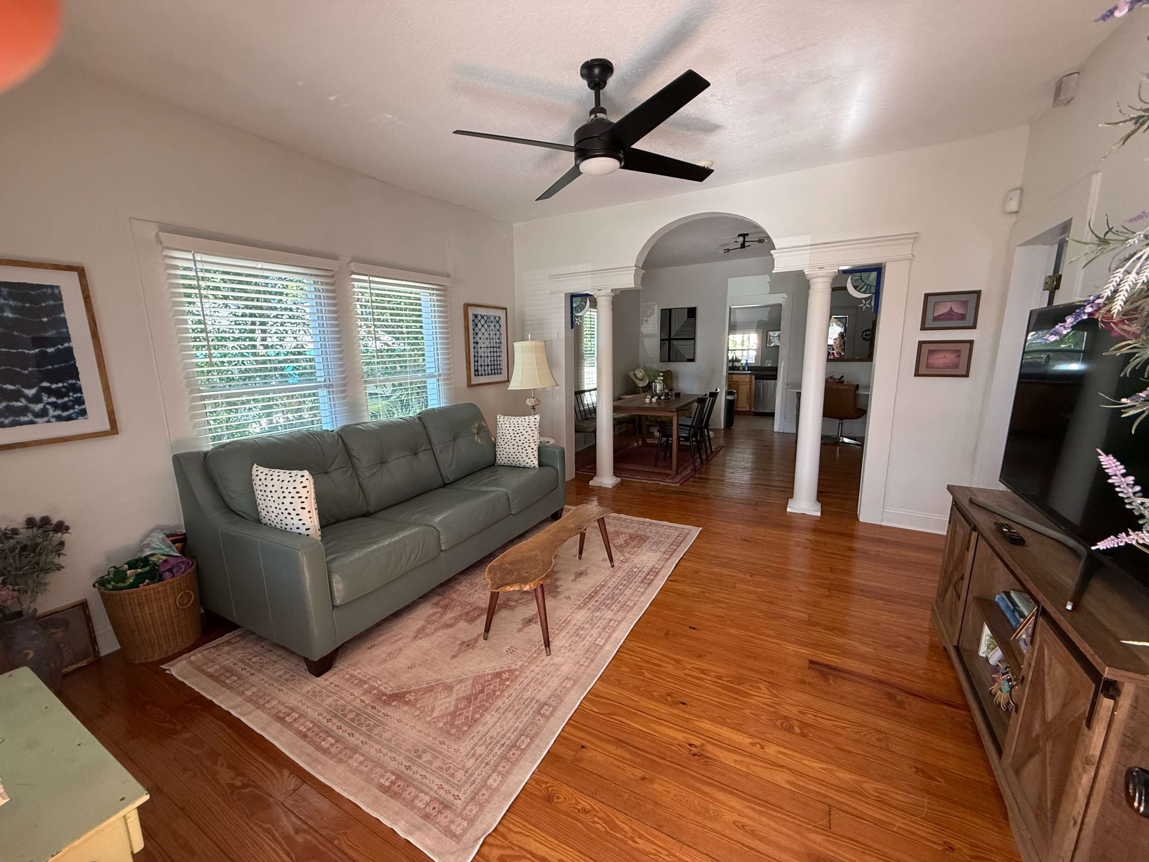 A living room features a green couch, a wooden coffee table, and a decorative rug, with archways leading to a dining area and kitchen in the background.