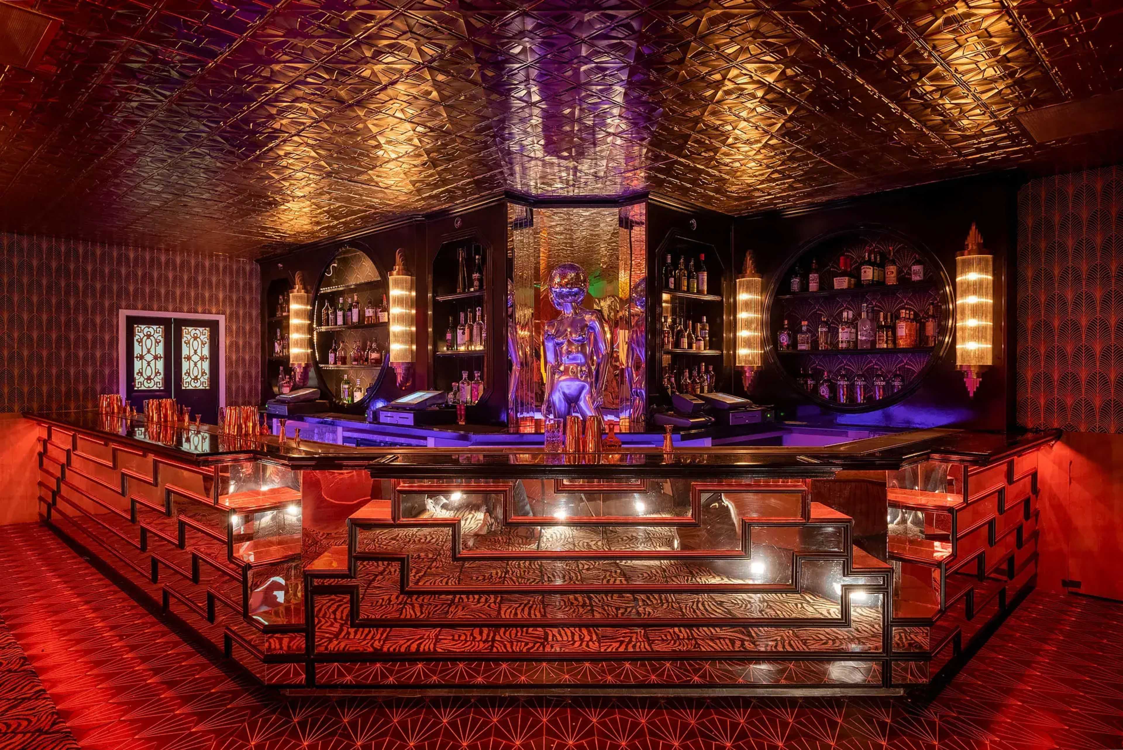 The image features an elegantly designed bar with a mirrored, stepped counter and shelves of liquor against a richly patterned backdrop.