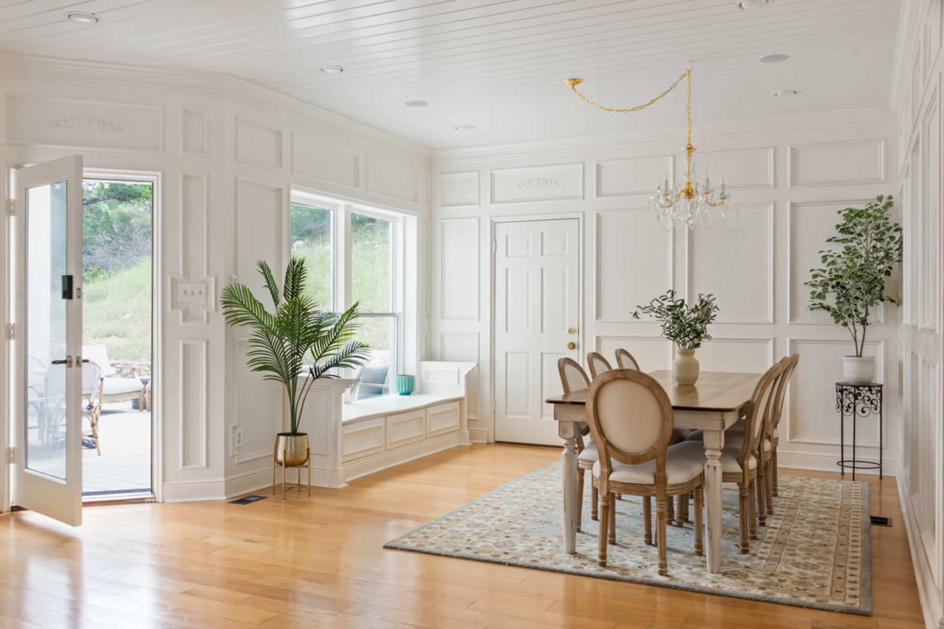 A bright dining area features a wooden table surrounded by upholstered chairs, with large windows allowing natural light to illuminate the space.
