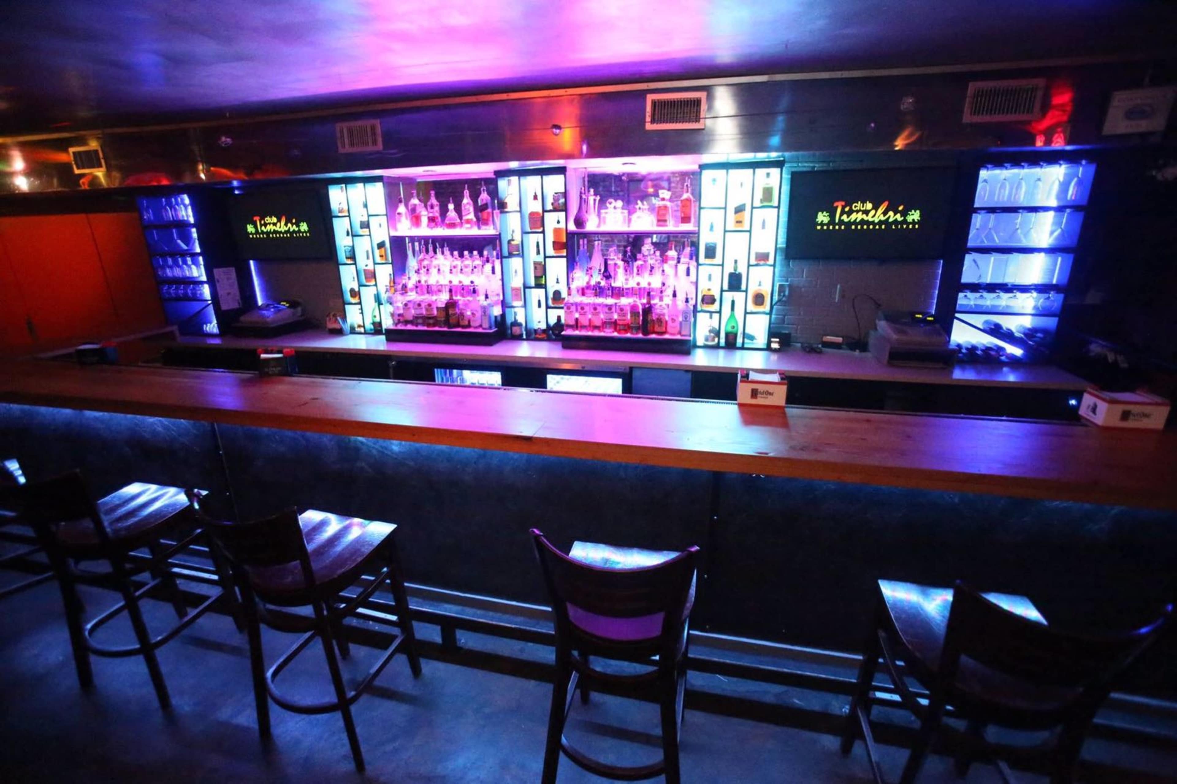 A dimly lit bar with a wooden counter and colorful illuminated shelves displaying various liquor bottles.
