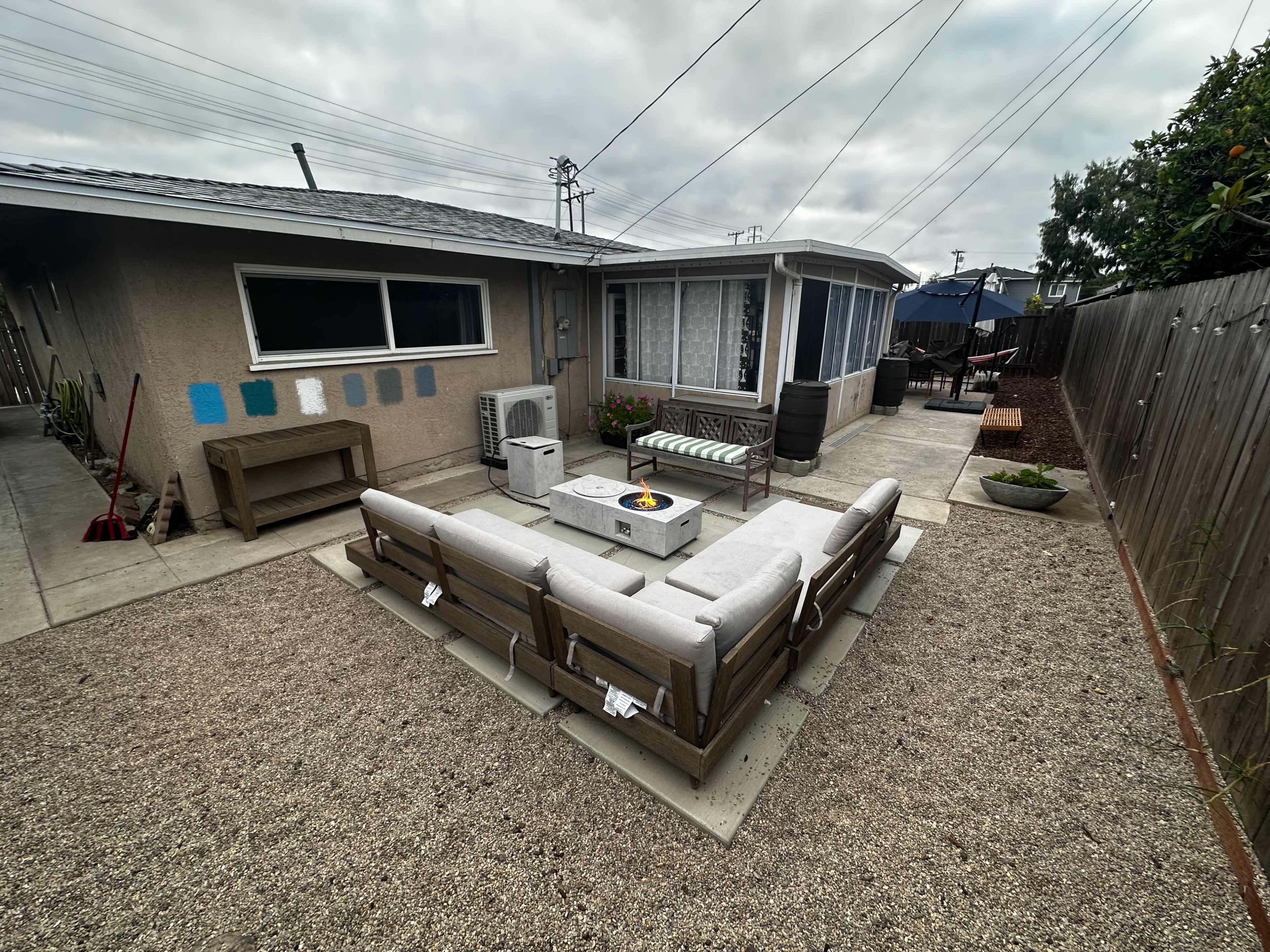 The image shows a backyard patio area featuring a seating arrangement around a fire pit, with a house and garden elements in the background.