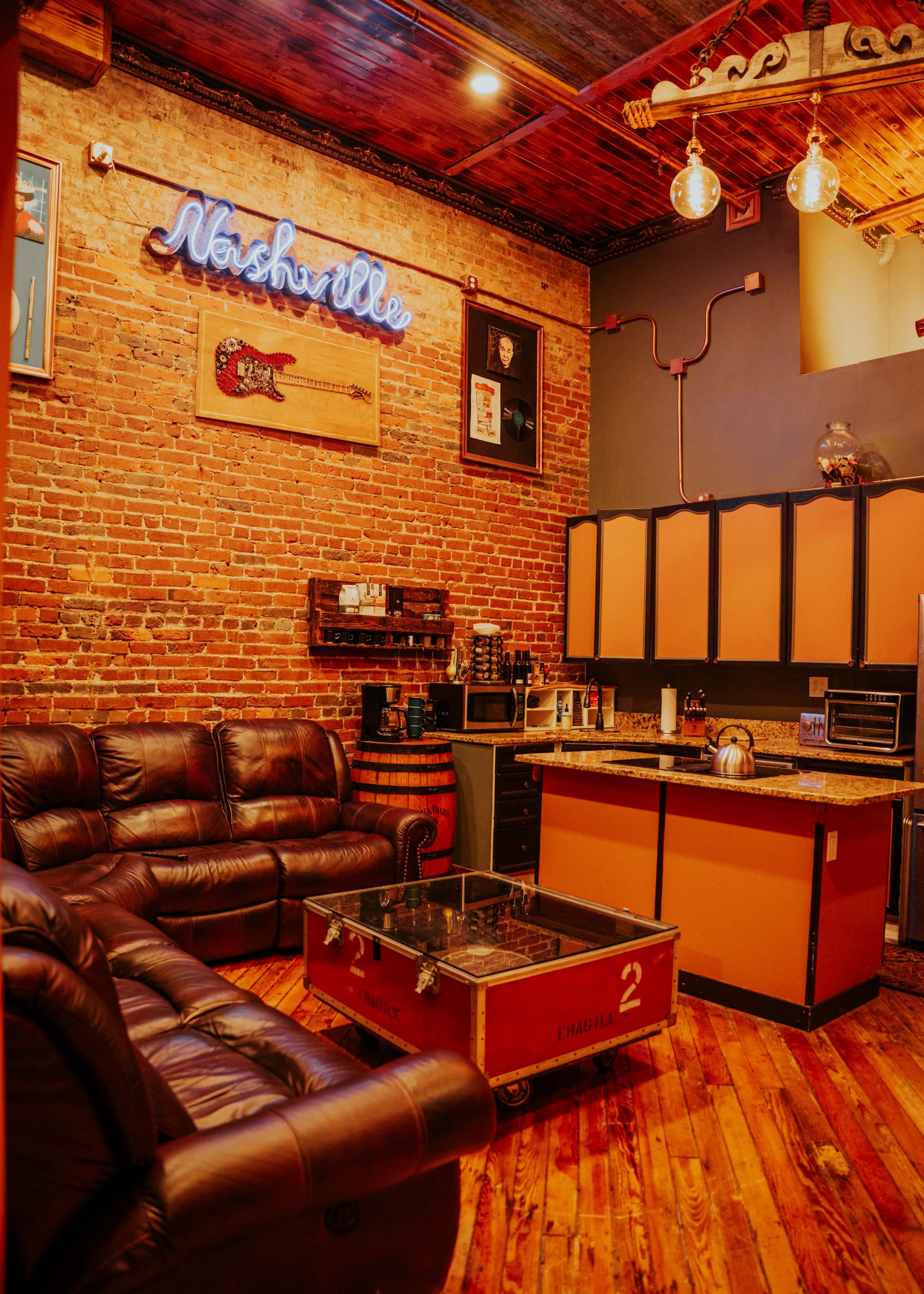 The image shows a cozy room with exposed brick walls, a dark leather couch, an orange kitchen area, and a wooden floor, complemented by a retro "Nashville" neon sign.