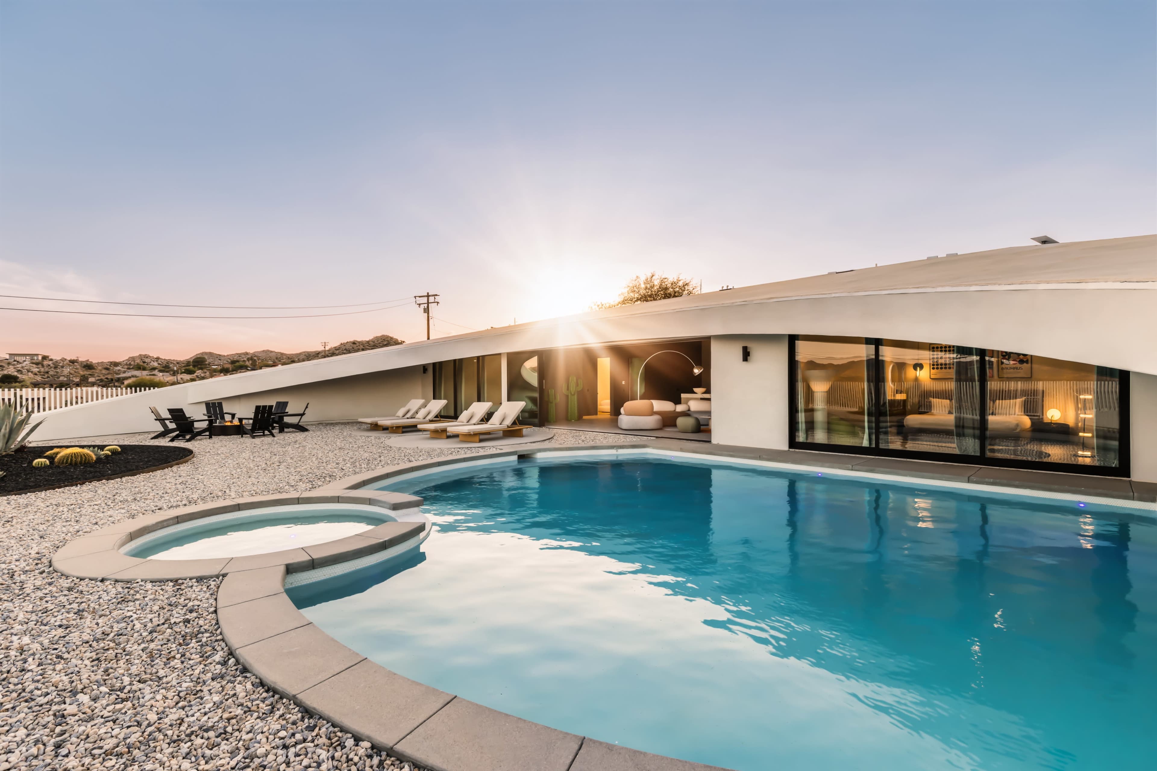 A modern home with large glass windows features a swimming pool and lounge chairs, set against a backdrop of a sunset.