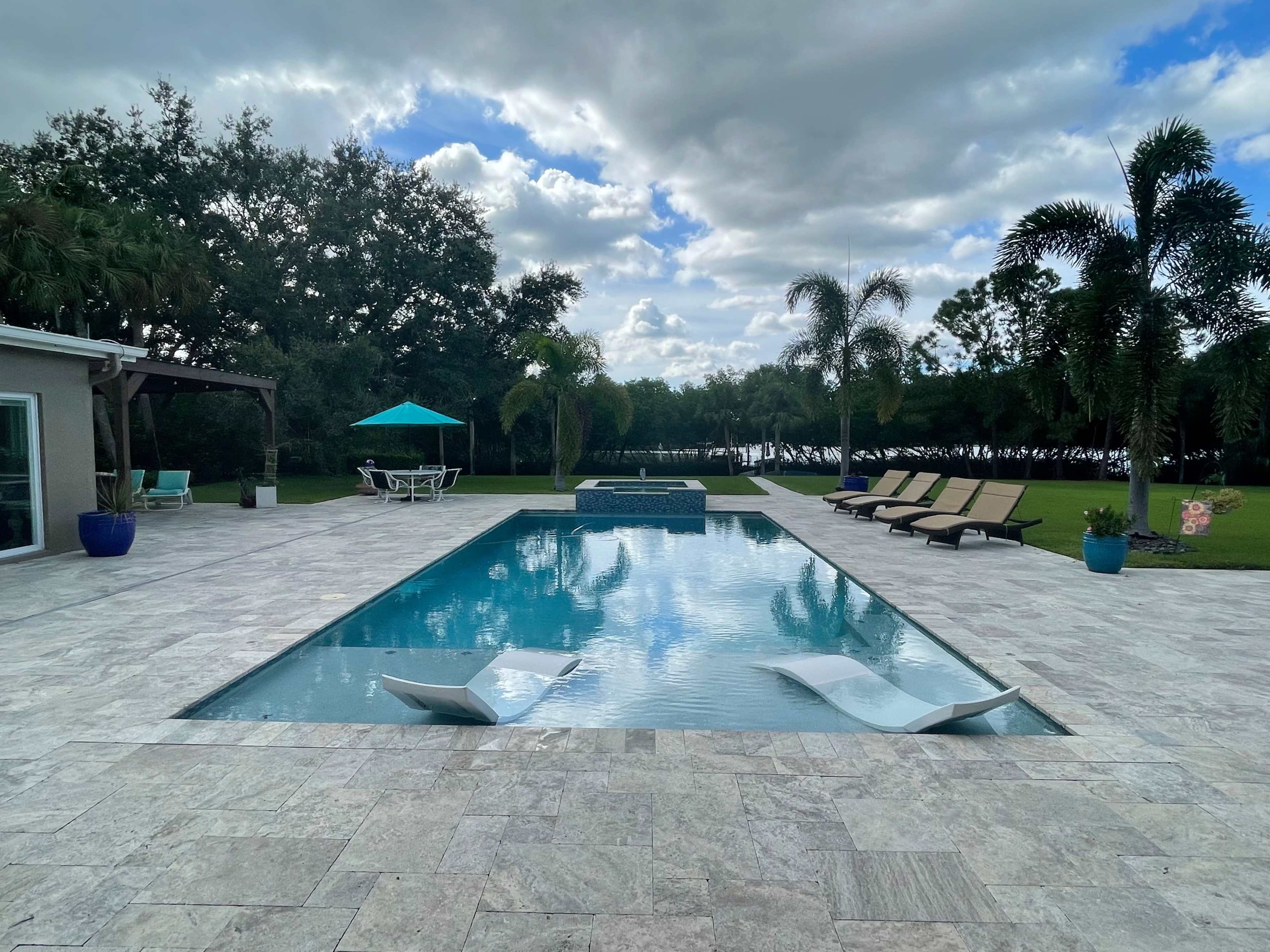 A rectangular pool surrounded by lounge chairs and a patio, with palm trees and green foliage in the background under a cloudy sky.