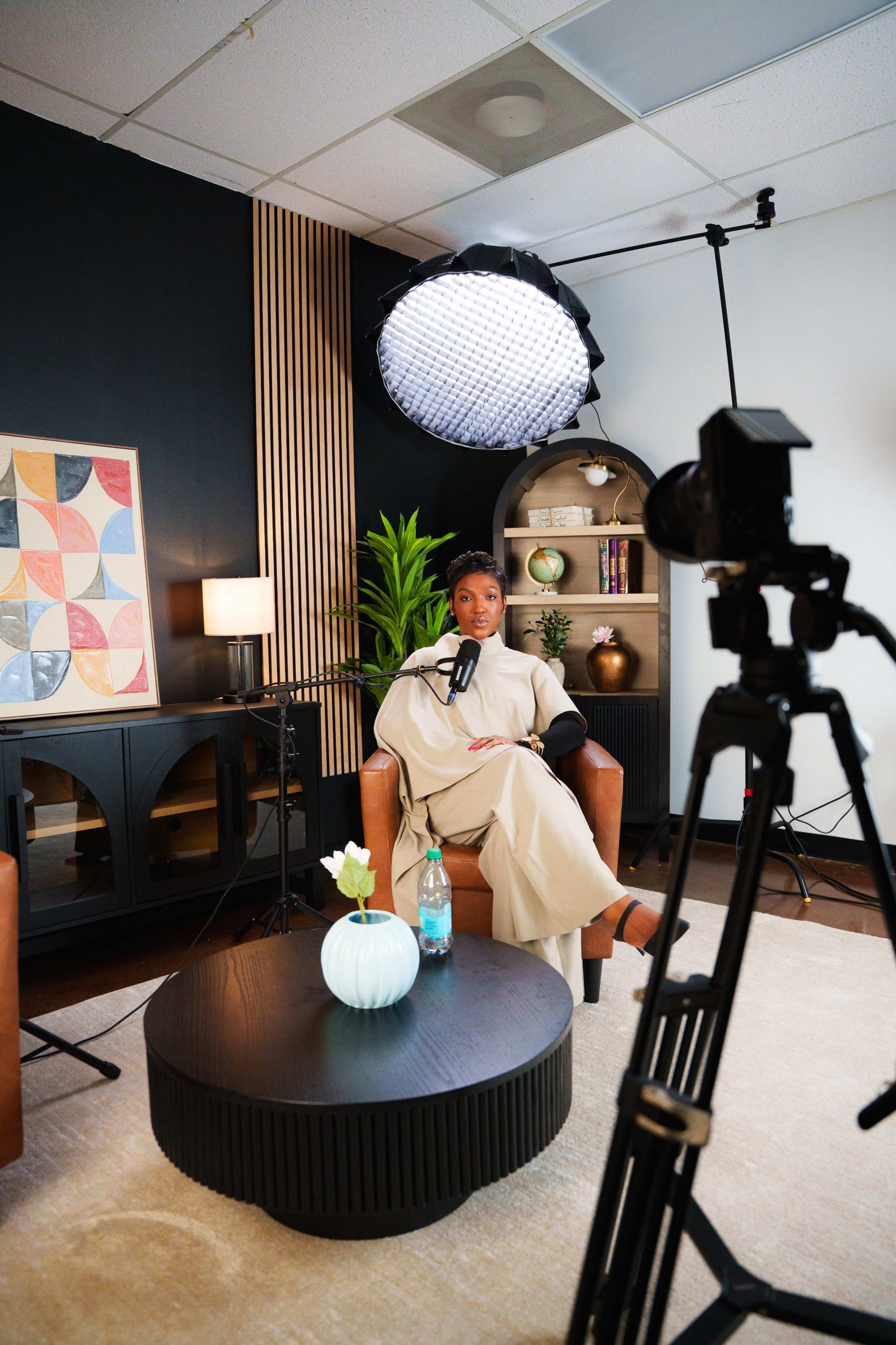 A woman sits in a studio setting with two brown armchairs, a black coffee table, and a backdrop of plants and decorative shelves, while a camera is positioned to record the scene.