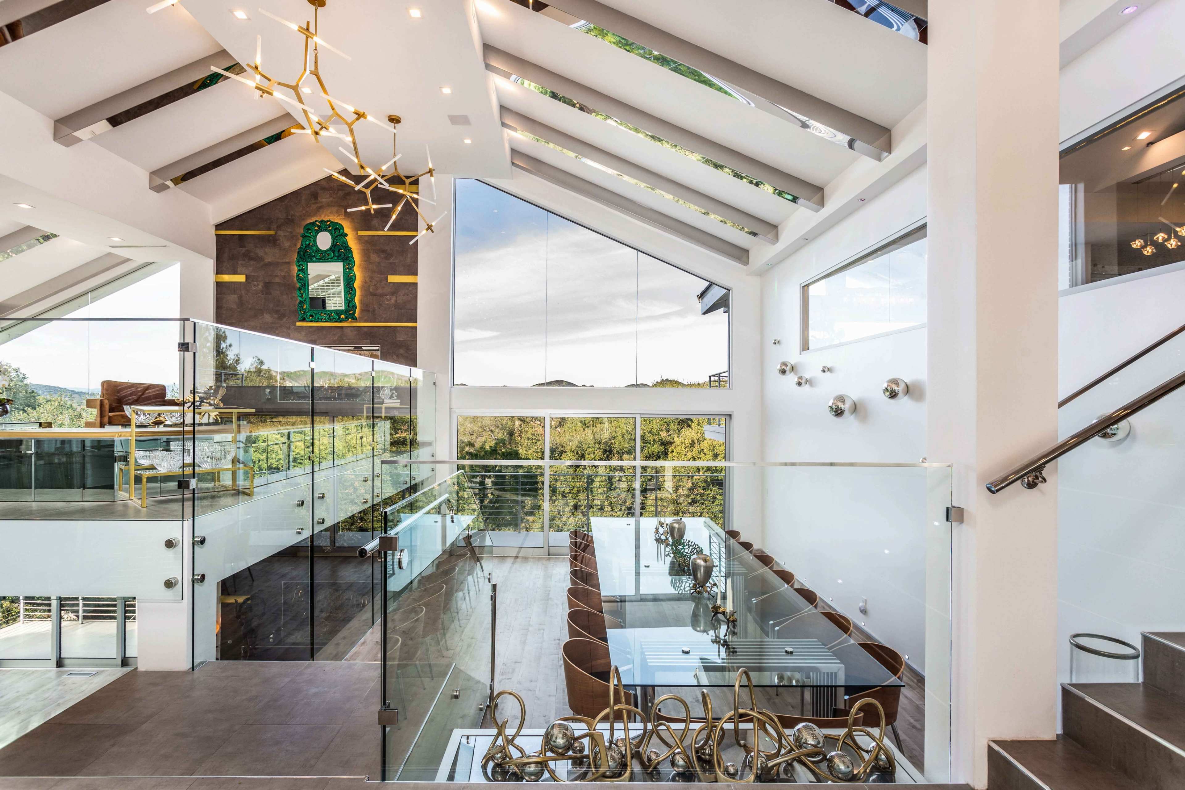 A modern interior space with a glass railing, a dining area featuring a long table, and large windows offering views of greenery.