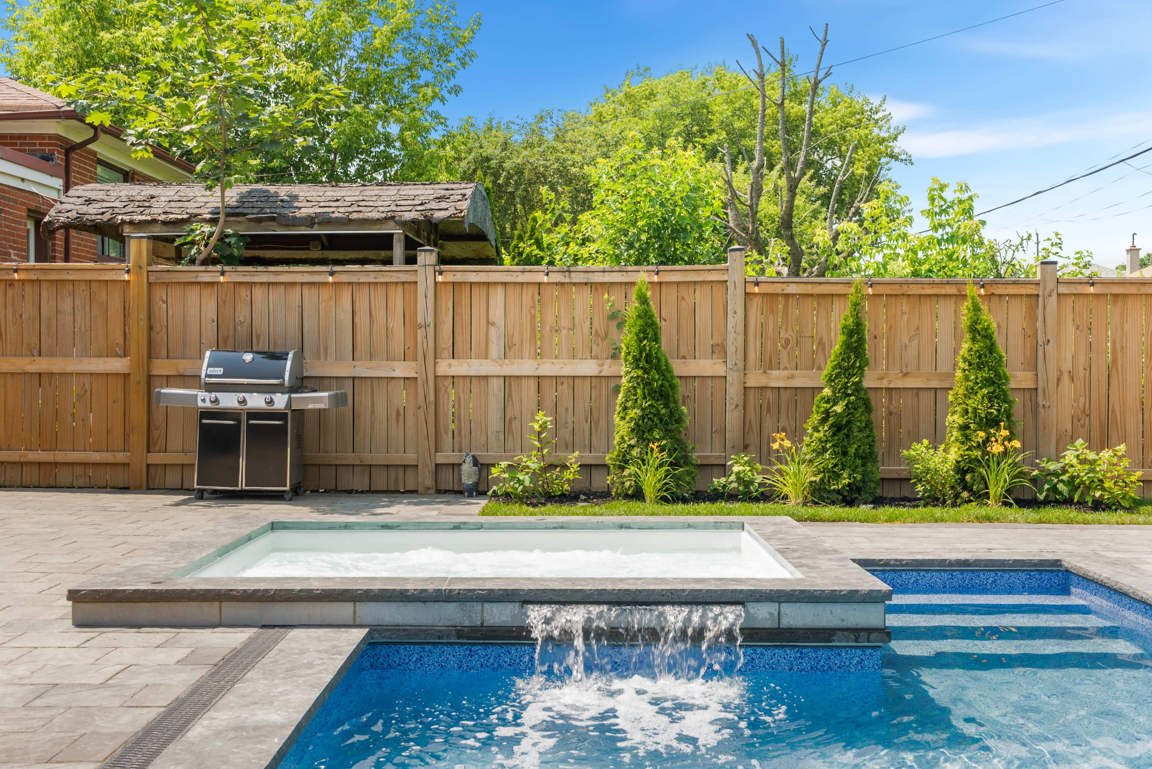 The image shows a backyard with a hot tub and swimming pool, accompanied by a barbecue grill and landscaped greenery behind wooden fencing.