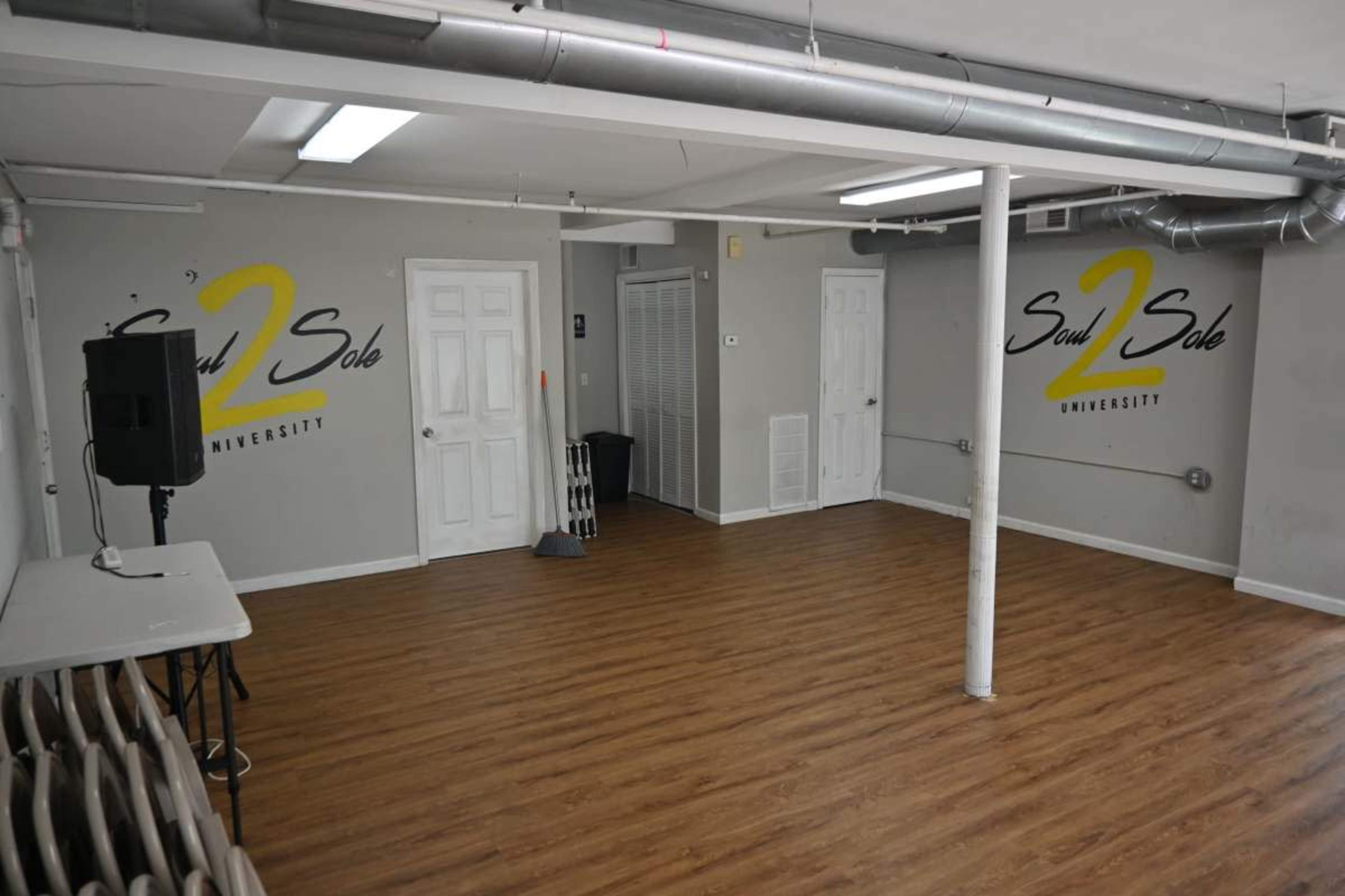 A spacious, well-lit room with wooden flooring, featuring a speaker system, a folding table, and the logo "Soul 2 Sole University" on the walls.