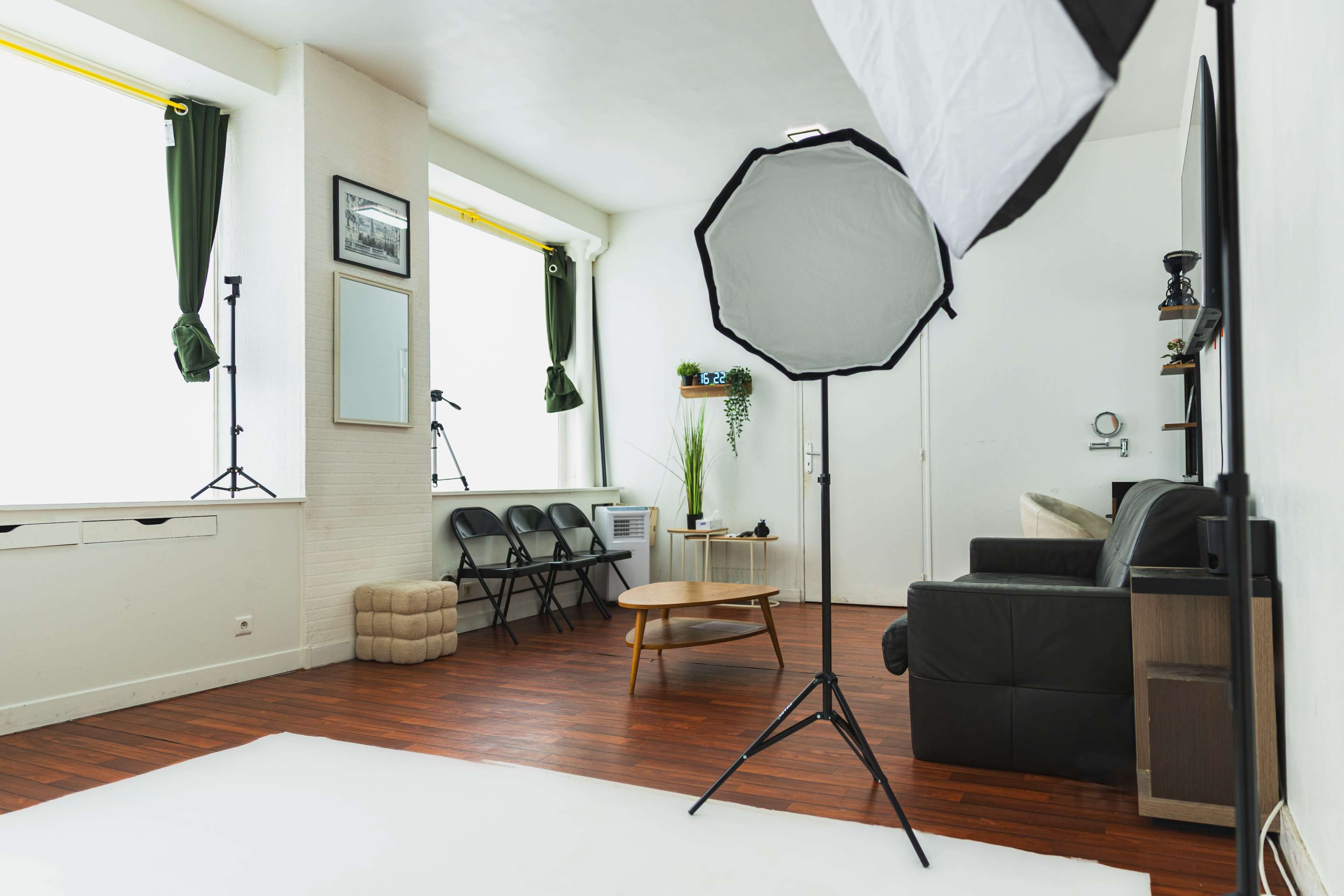 The image shows a well-lit photography studio with a wooden floor, featuring a black leather couch, a table, and various props for photo shoots.