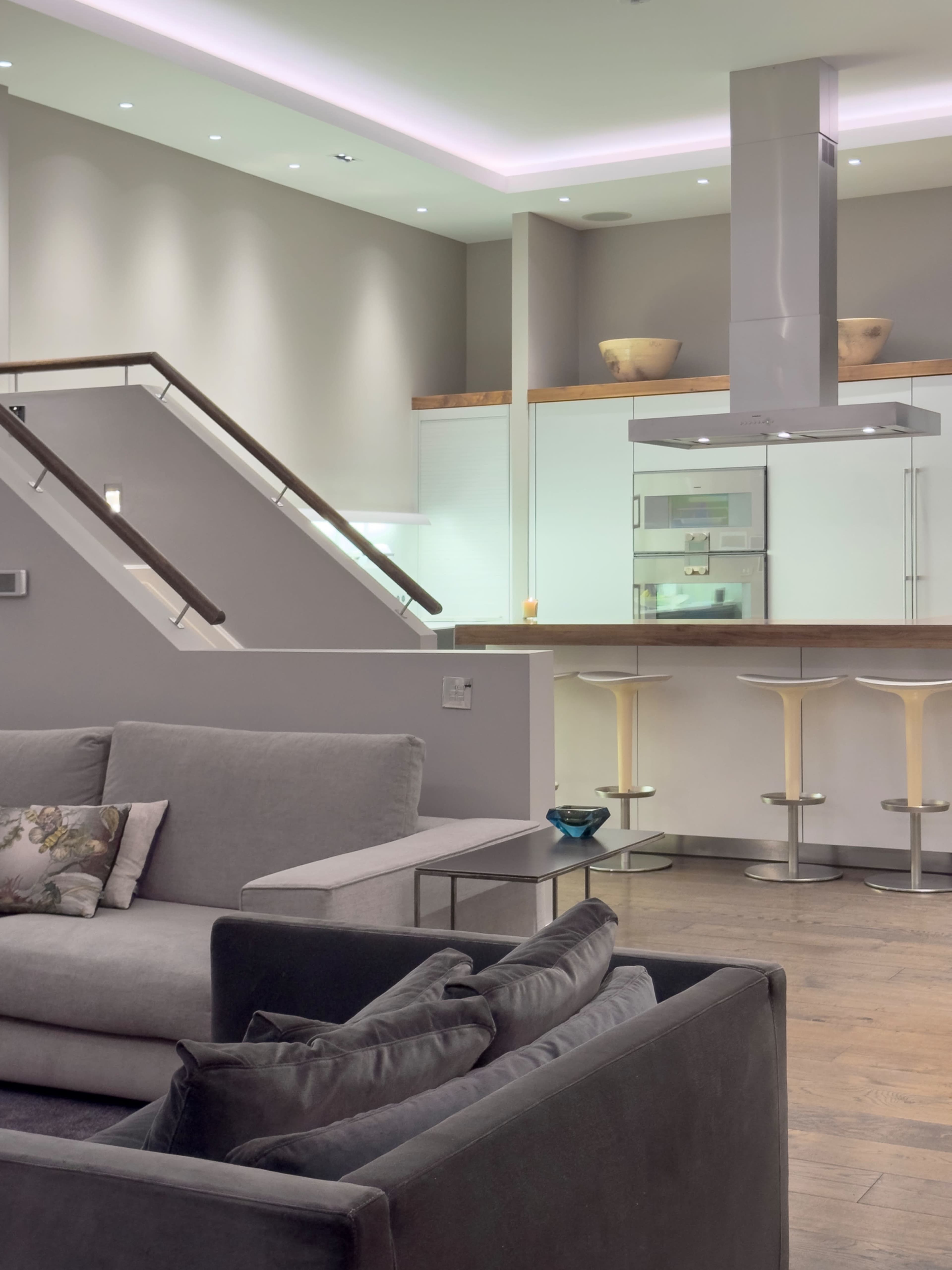 The image shows a modern kitchen and living area, featuring a staircase, a sofa, and a kitchen island with bar stools.