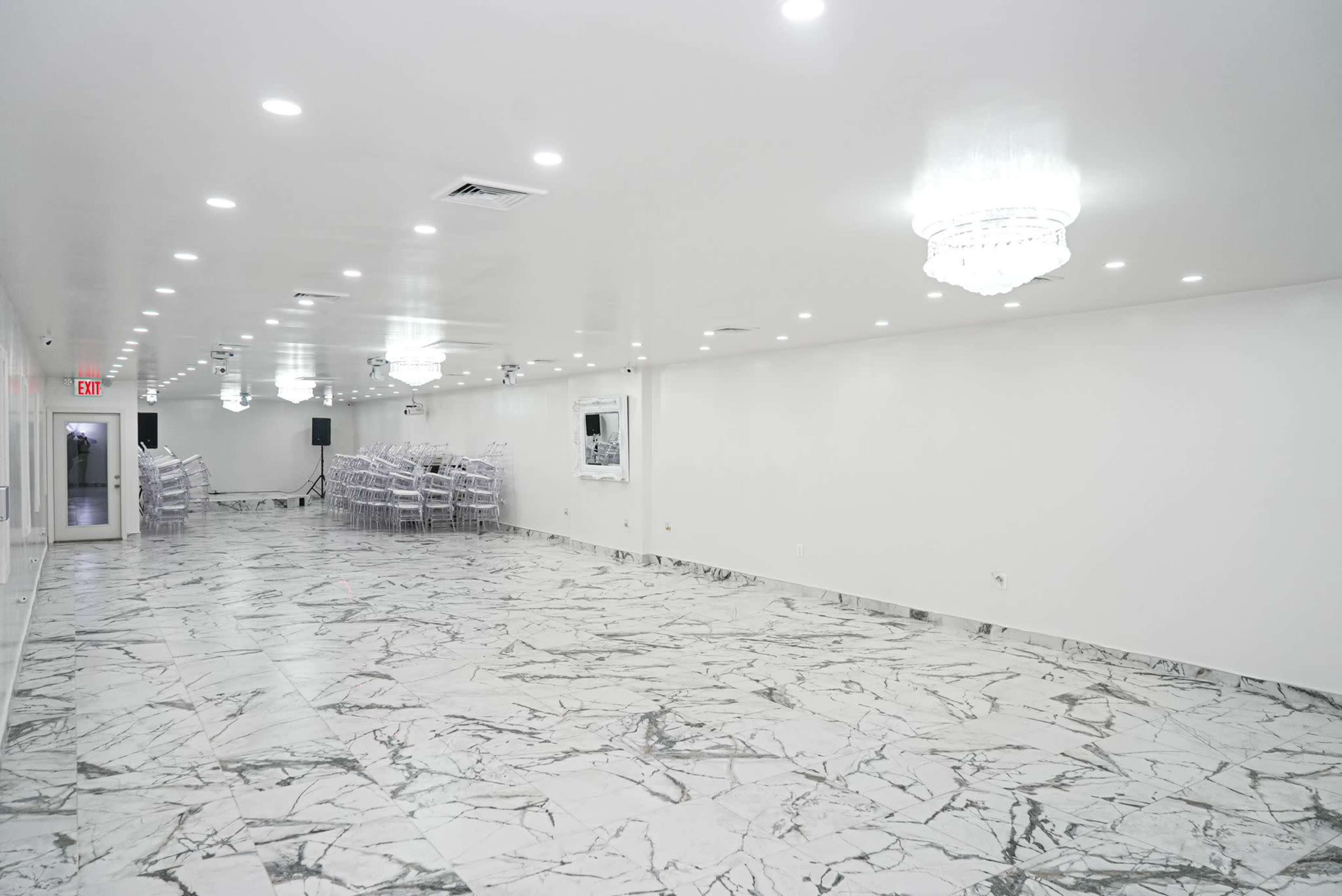 A spacious, well-lit hall features marble flooring and elegant chandeliers, with white walls and a row of stackable chairs along one side.