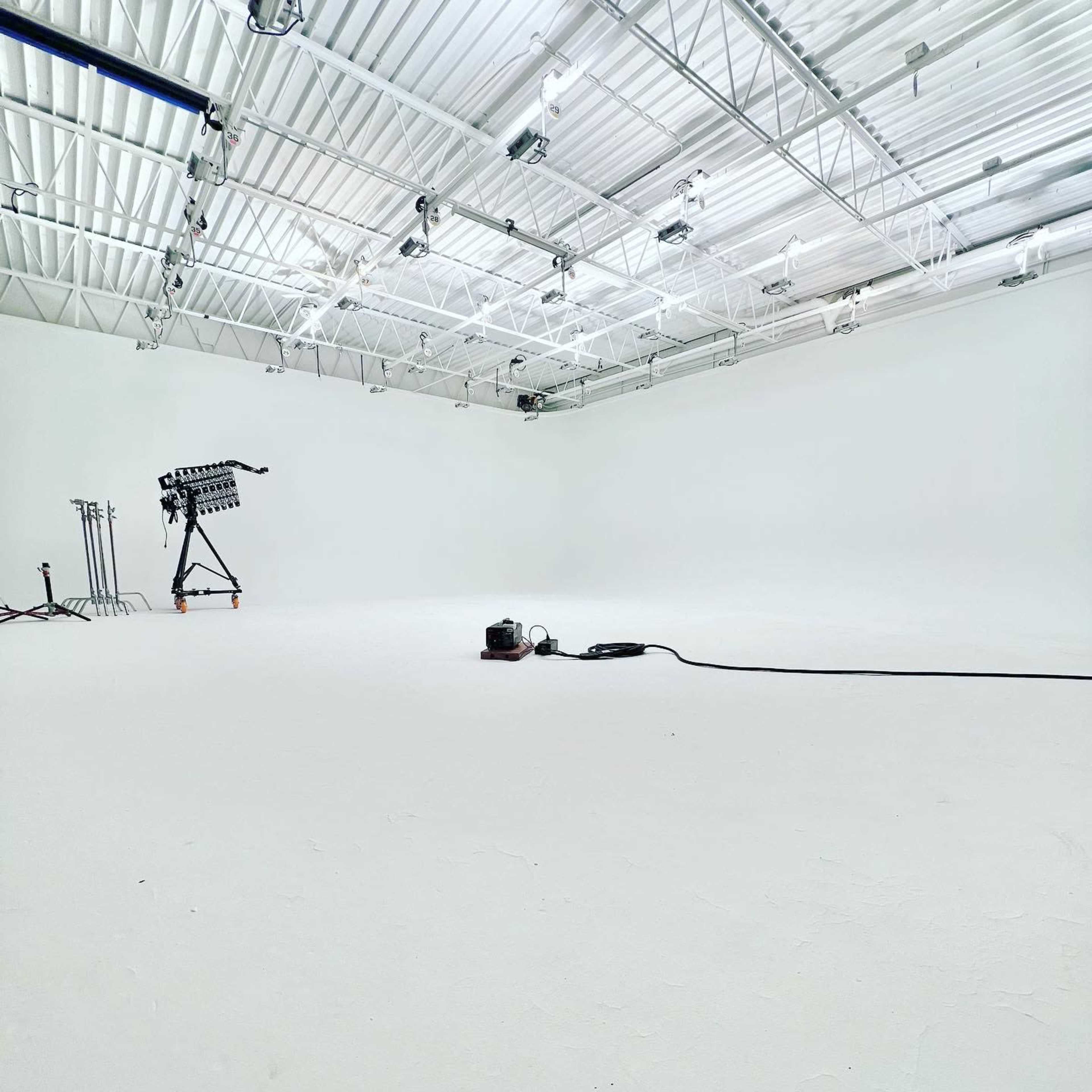 A spacious, empty studio with a white floor and walls, featuring lighting equipment on stands and a vacuum cleaner on the floor.