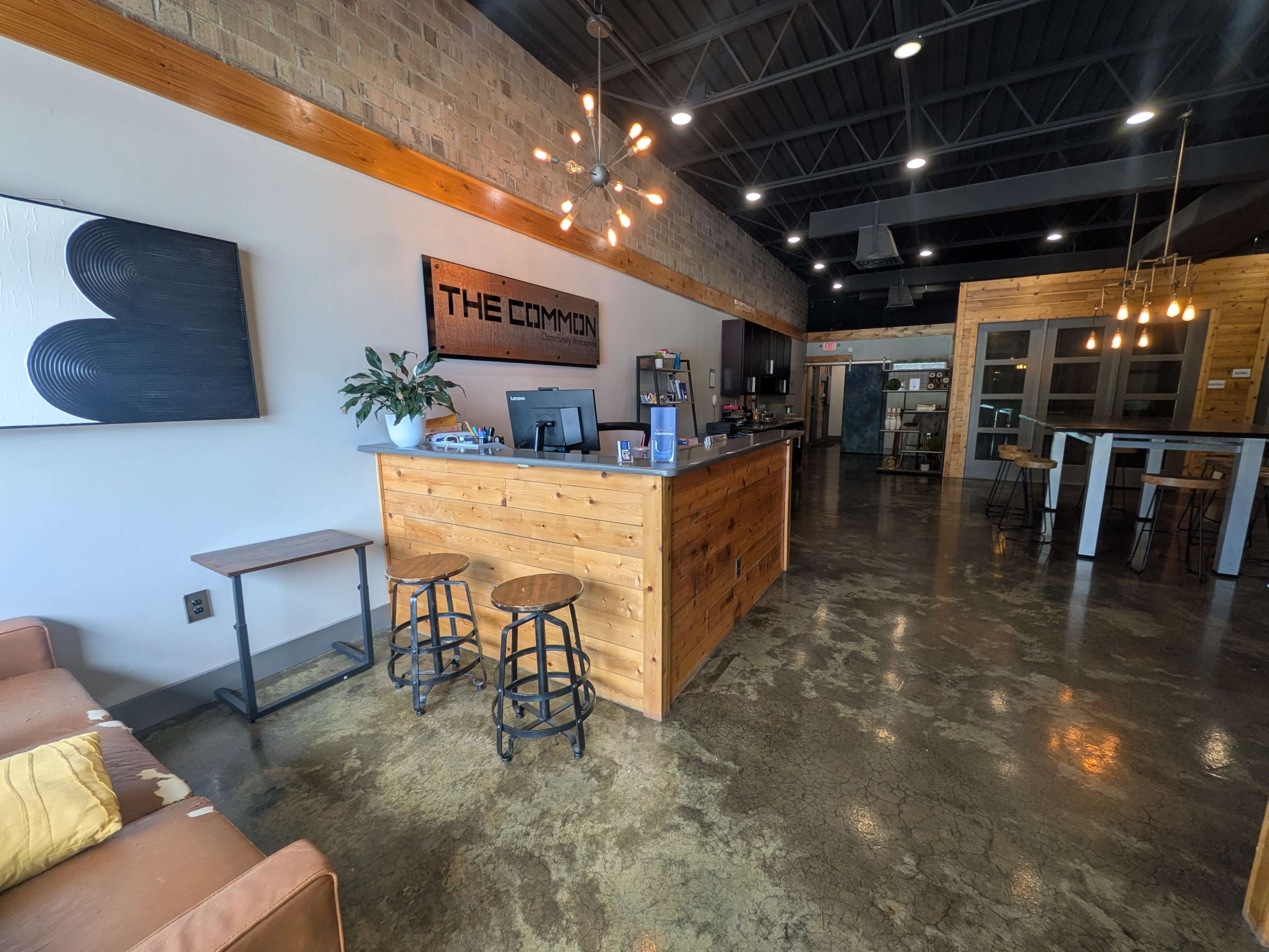 The image shows a cozy, modern interior of a café or communal space with a wooden counter, seating area, and industrial-style lighting.