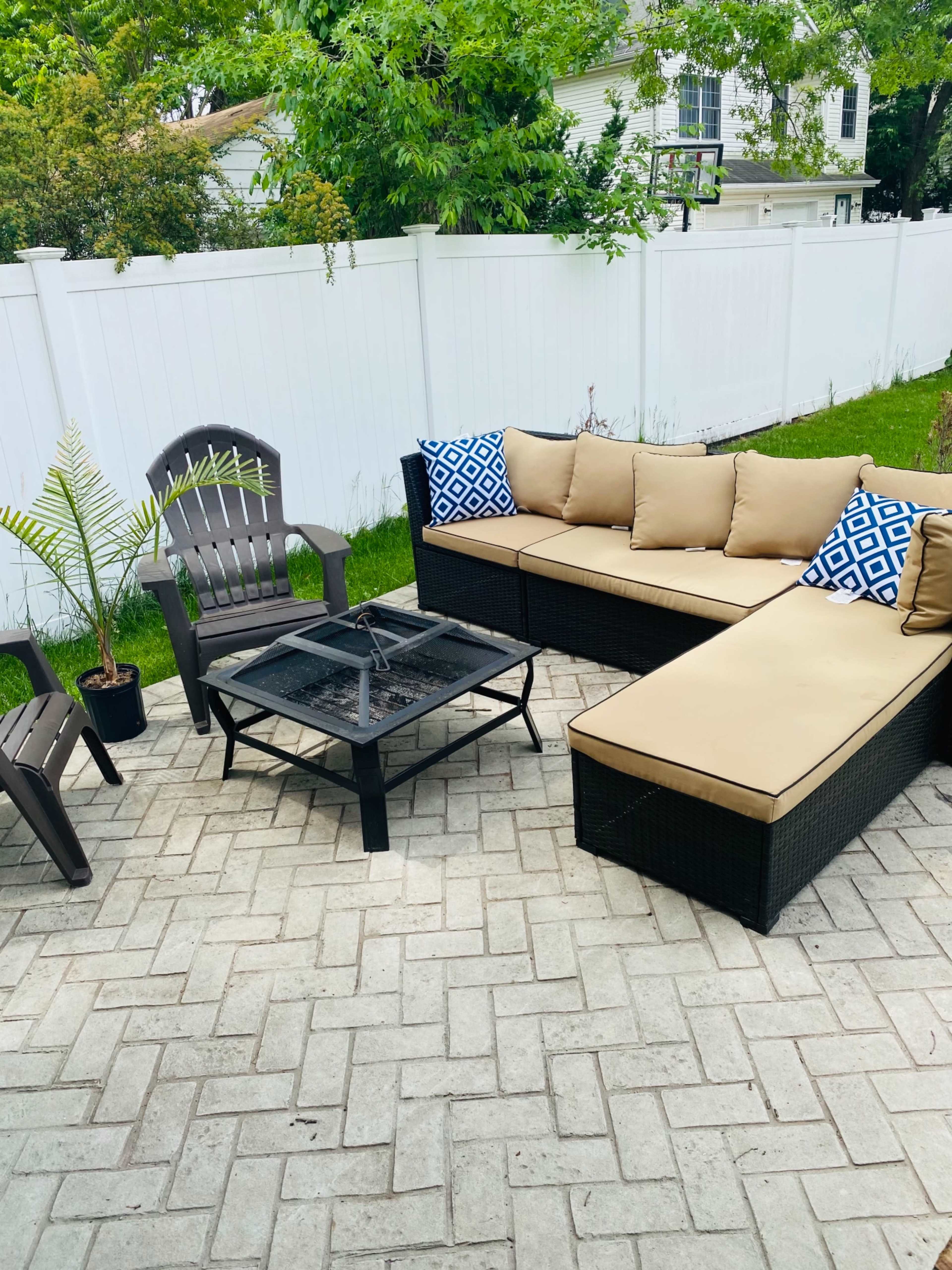 A patio area features a sectional sofa with beige cushions, a fire pit table, and two chairs, surrounded by a white fence and greenery.
