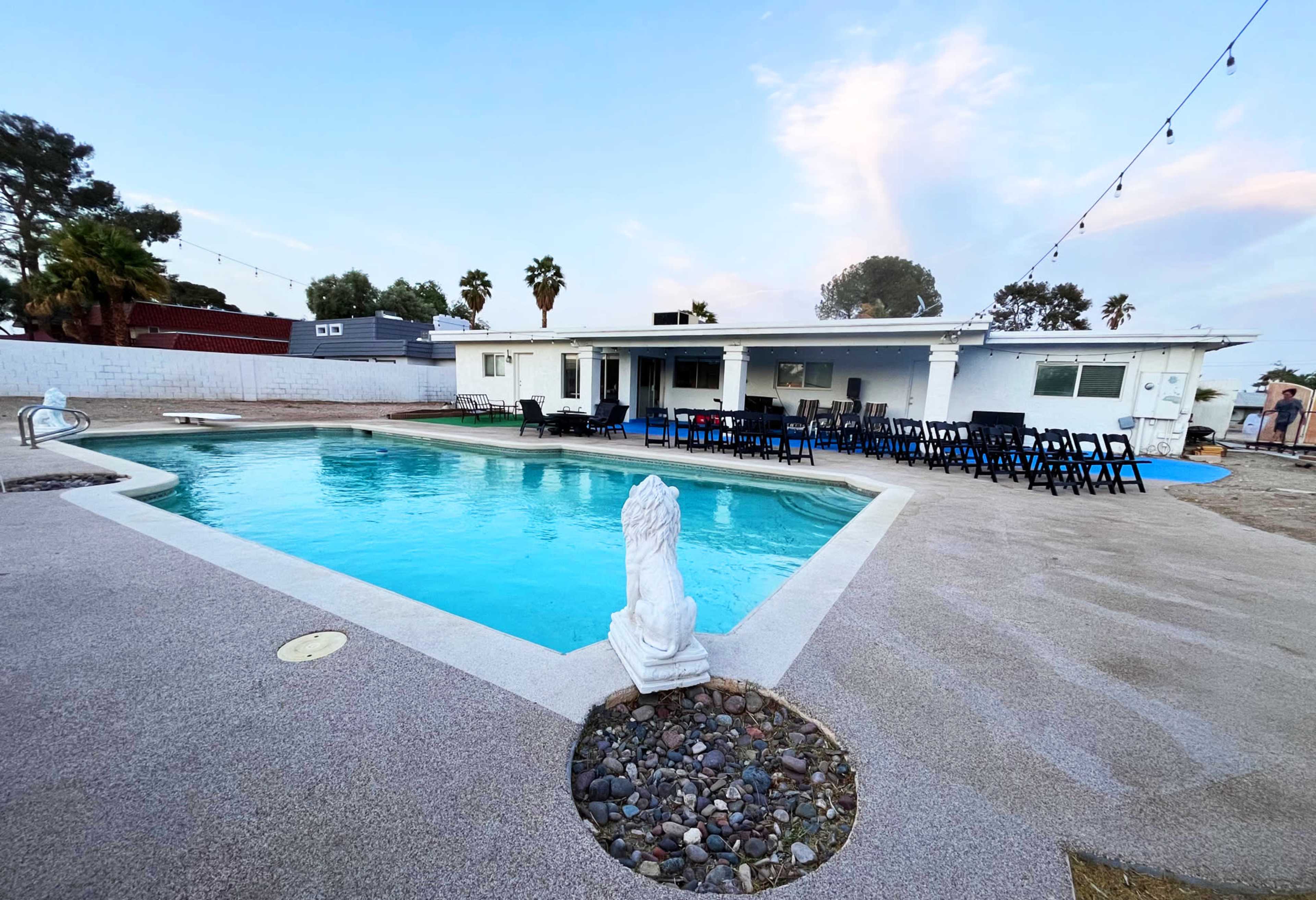 The image shows a swimming pool area with a white house in the background and several black chairs arranged around the pool.