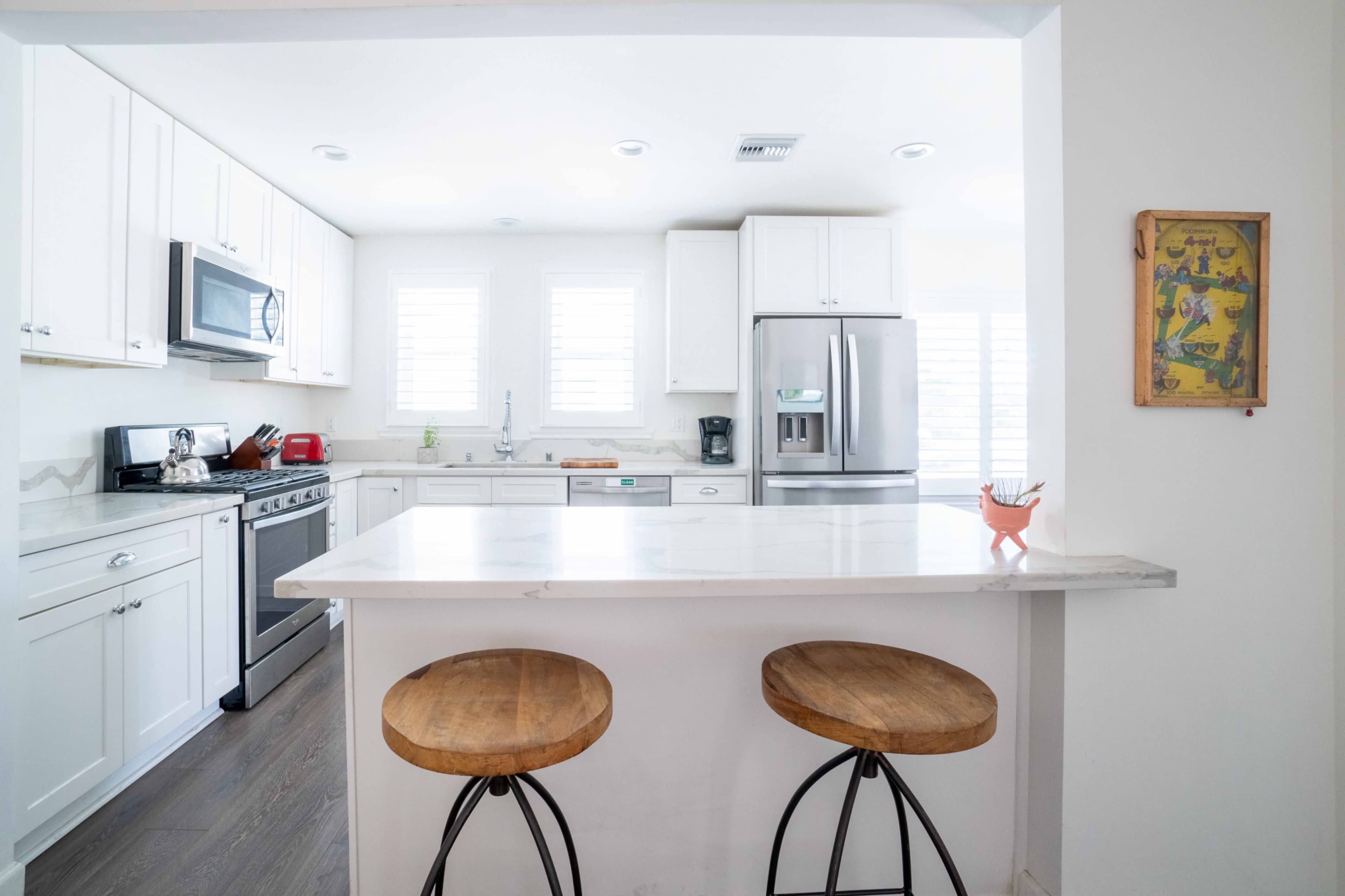 A bright modern kitchen features white cabinetry, stainless steel appliances, and a marble countertop with two wooden stools at the bar.