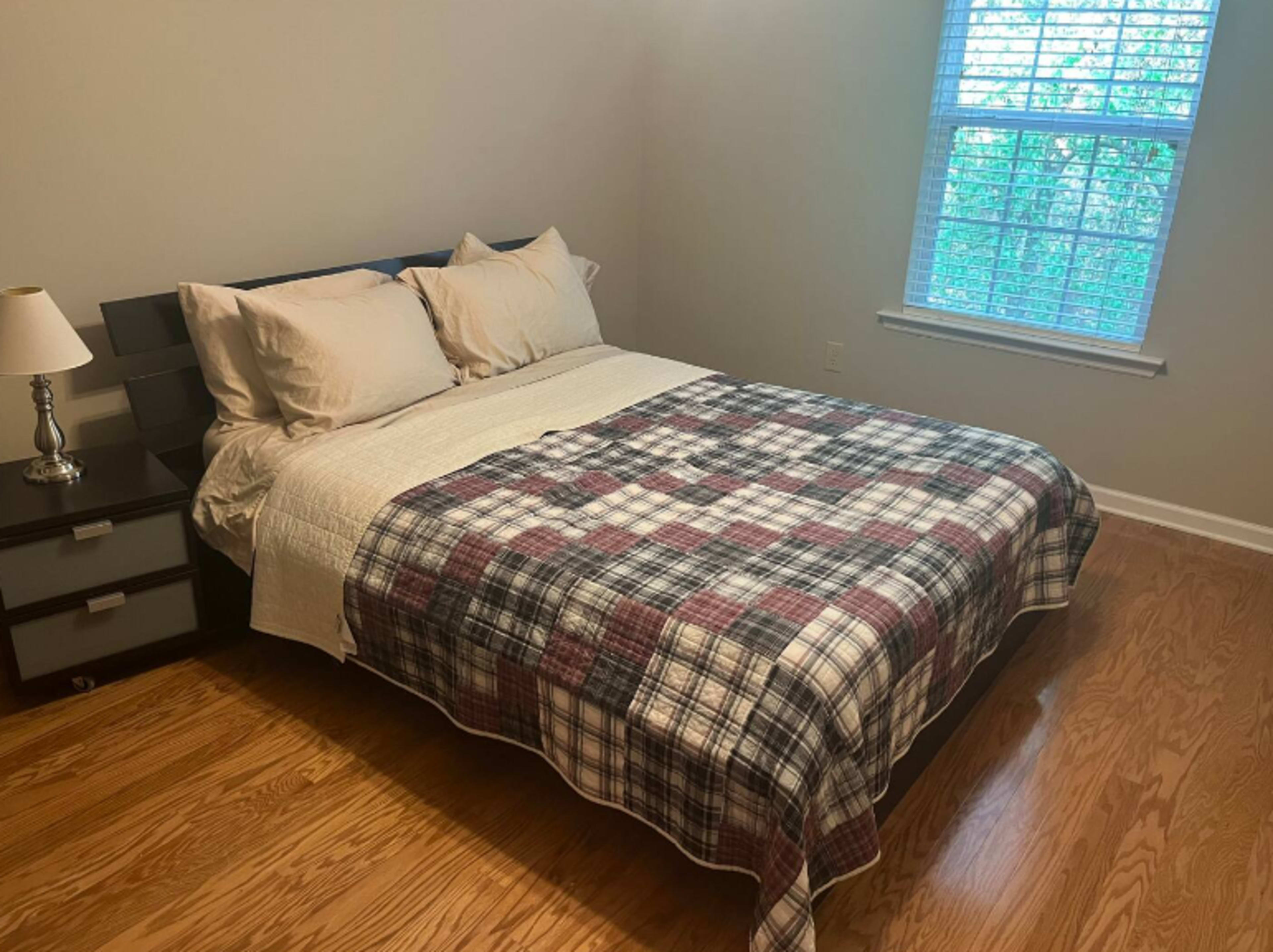 A neatly made bed with a checkered blanket is positioned in a softly lit bedroom beside a bedside lamp and a window.