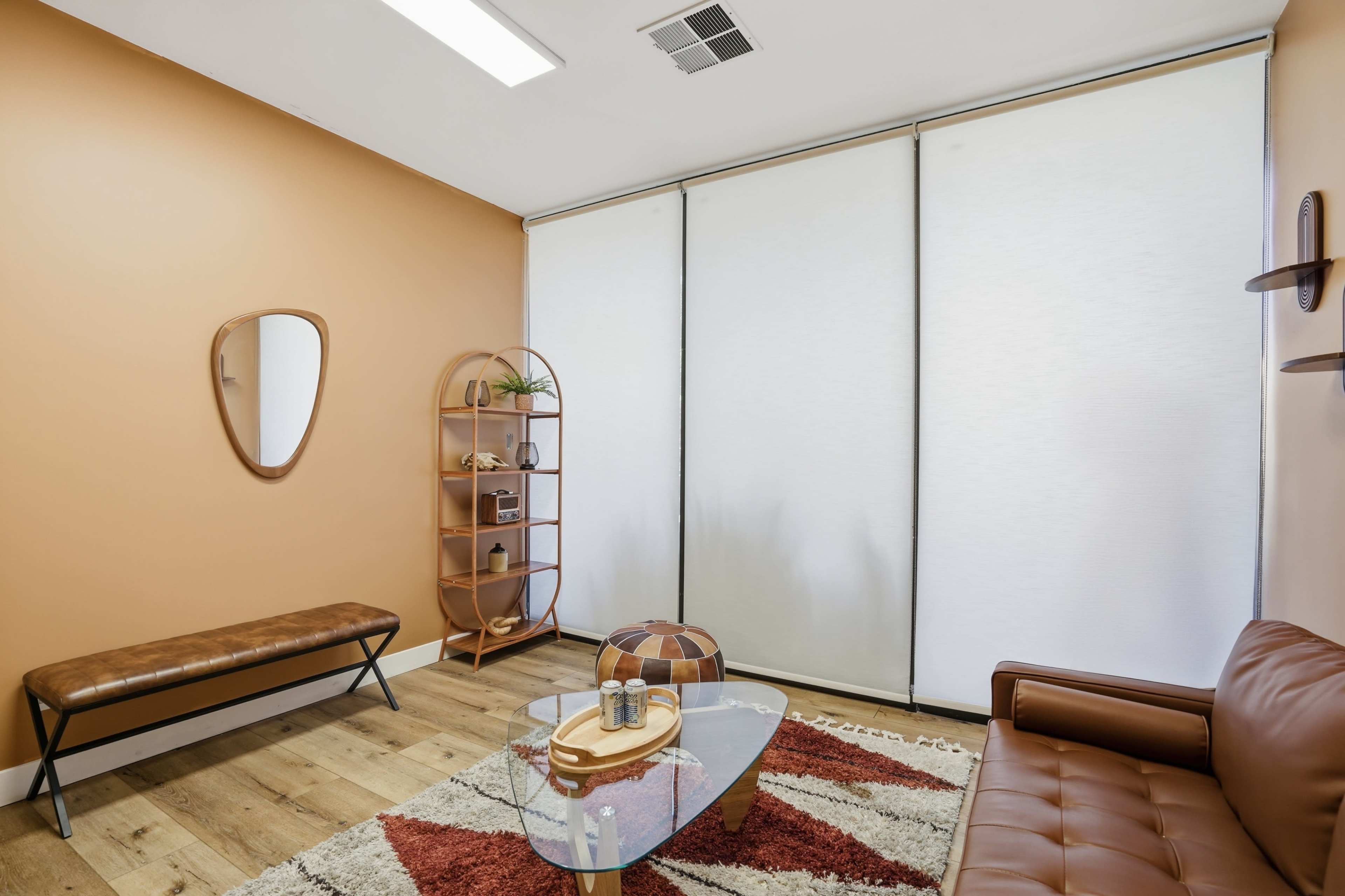 The image shows a small room with an orange wall, a brown leather sofa, a glass coffee table, a wooden bench, and a shelf with decorative items.