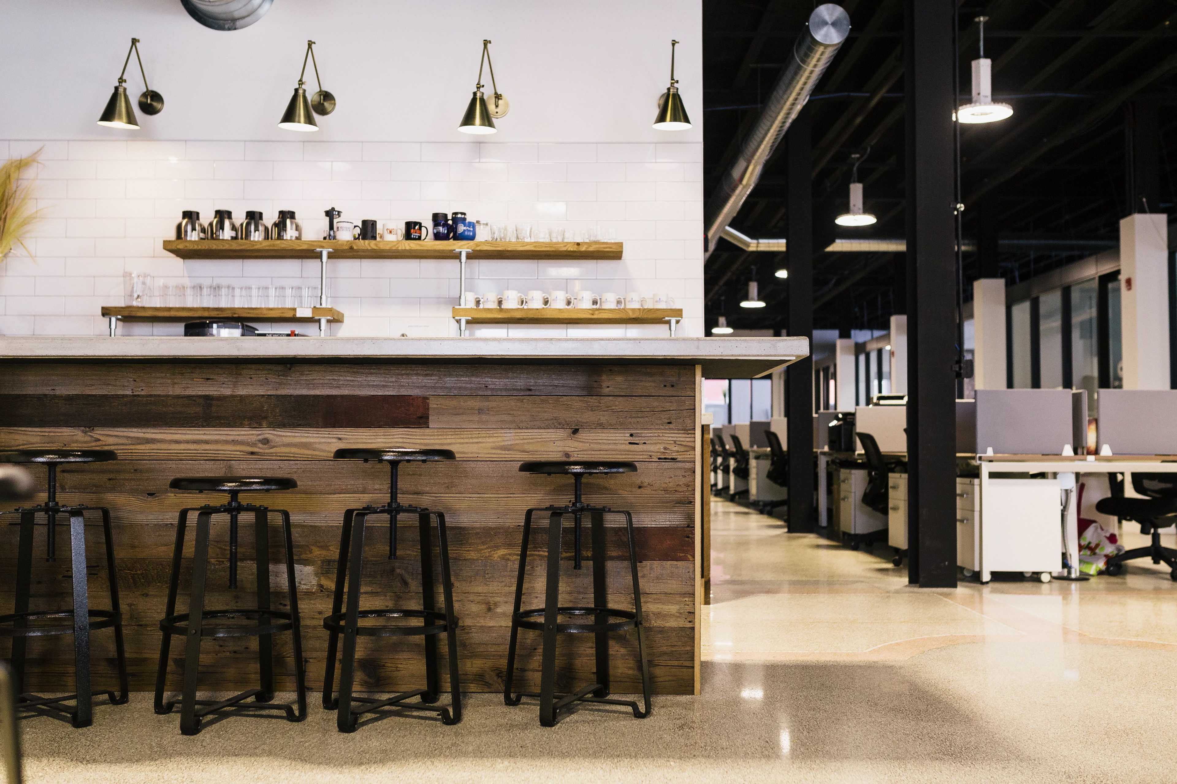 A modern office space features a wooden bar with high stools and a sleek countertop, behind which are shelves displaying coffee mugs and glassware.