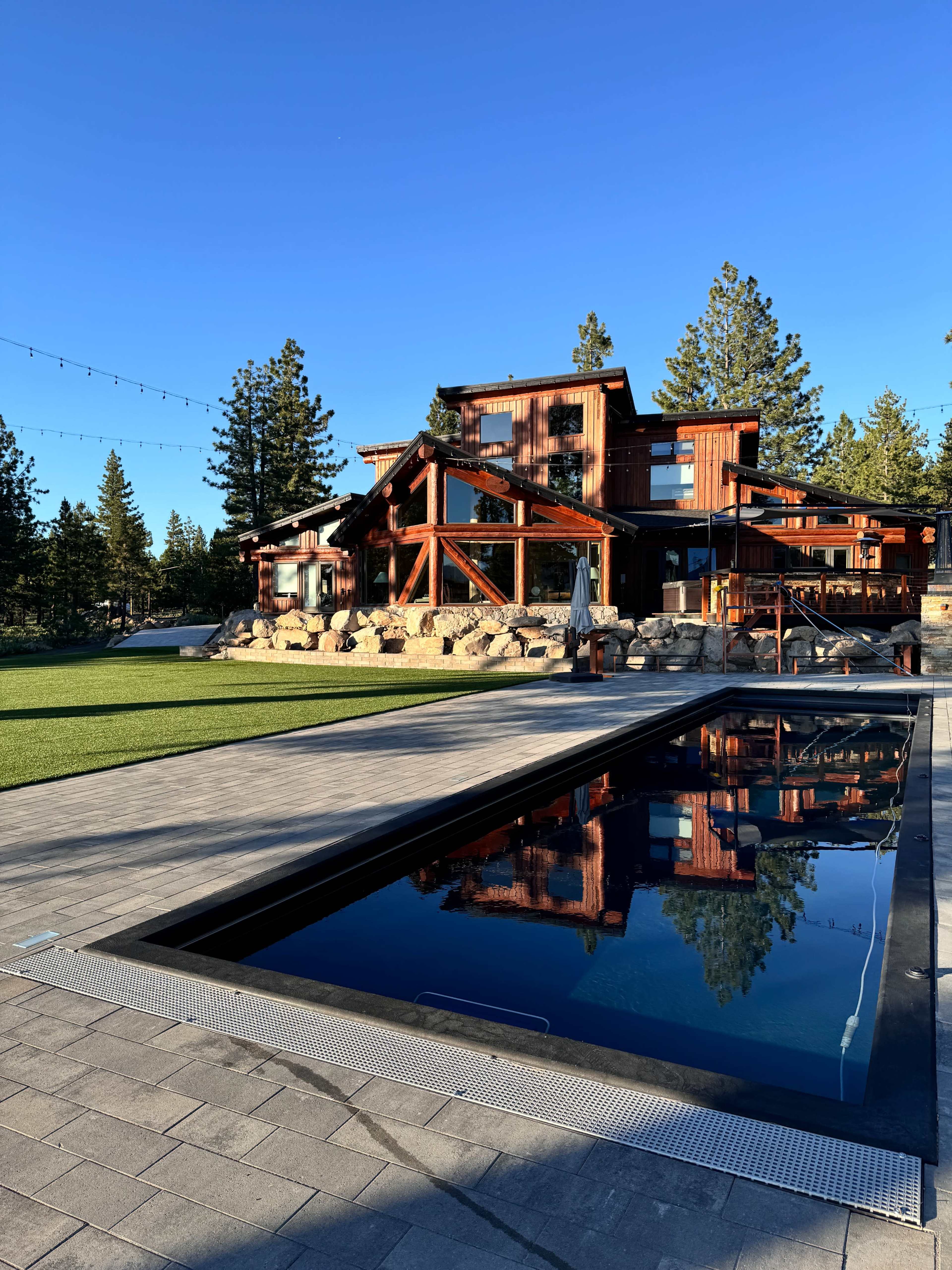A modern wooden house with large windows is set against a clear blue sky, reflecting in a sleek swimming pool in the foreground.