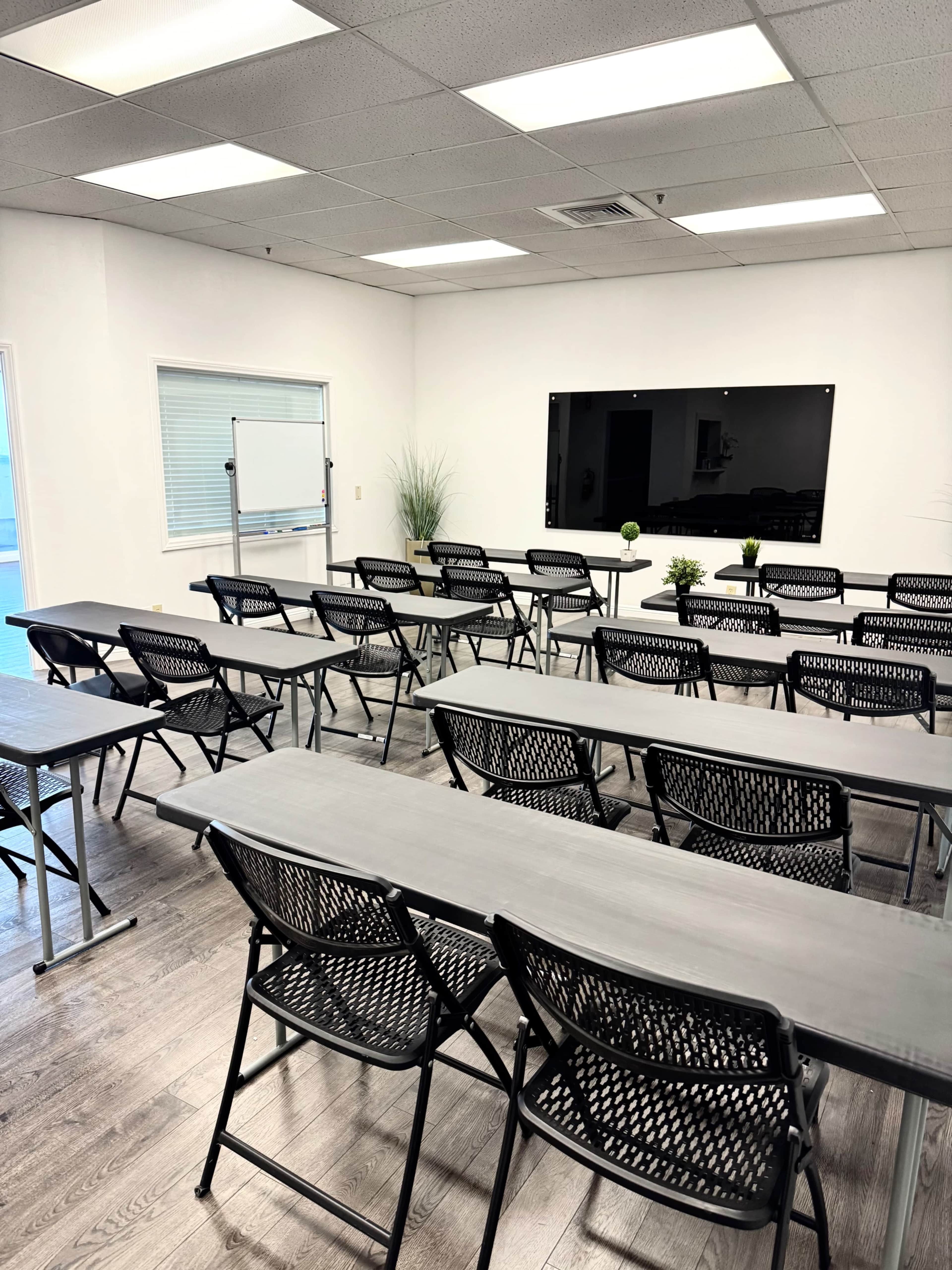 The image shows a classroom or meeting room arranged with rows of folding chairs and long tables, featuring a large black display screen on the wall and plants in the corners.