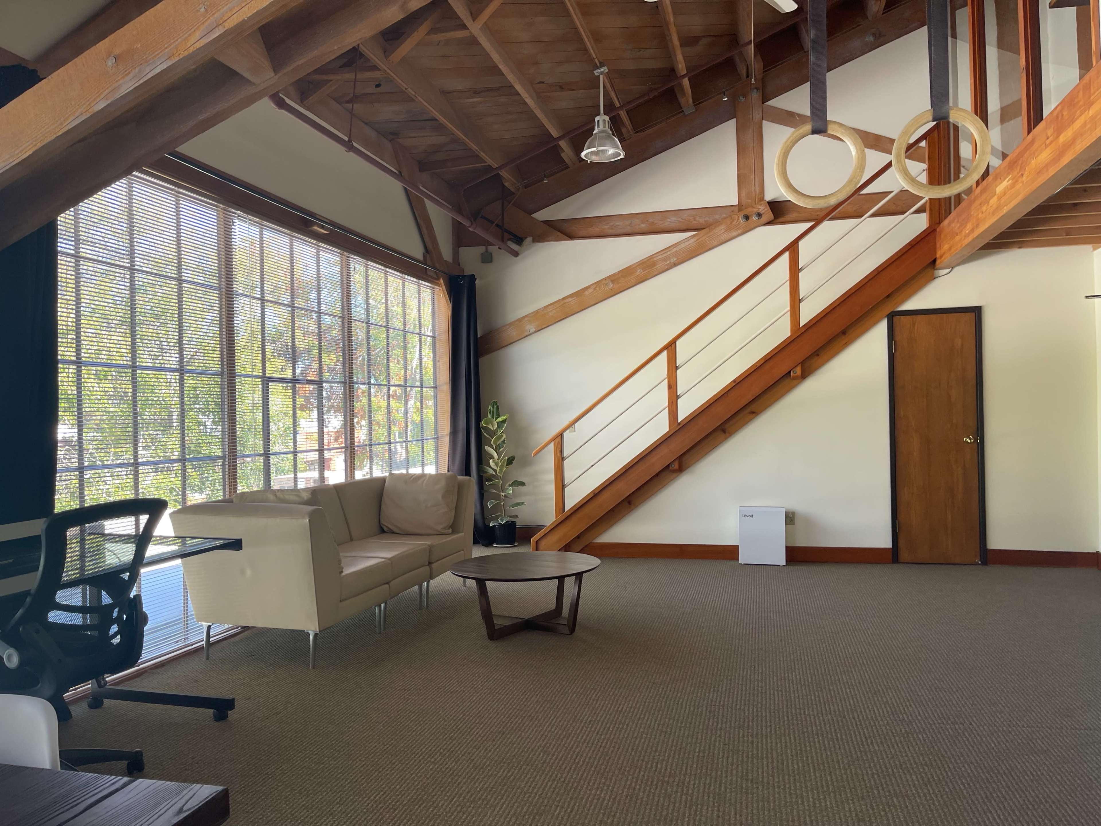 A spacious office interior featuring a staircase, a couch, a coffee table, and large windows allowing natural light to enter.