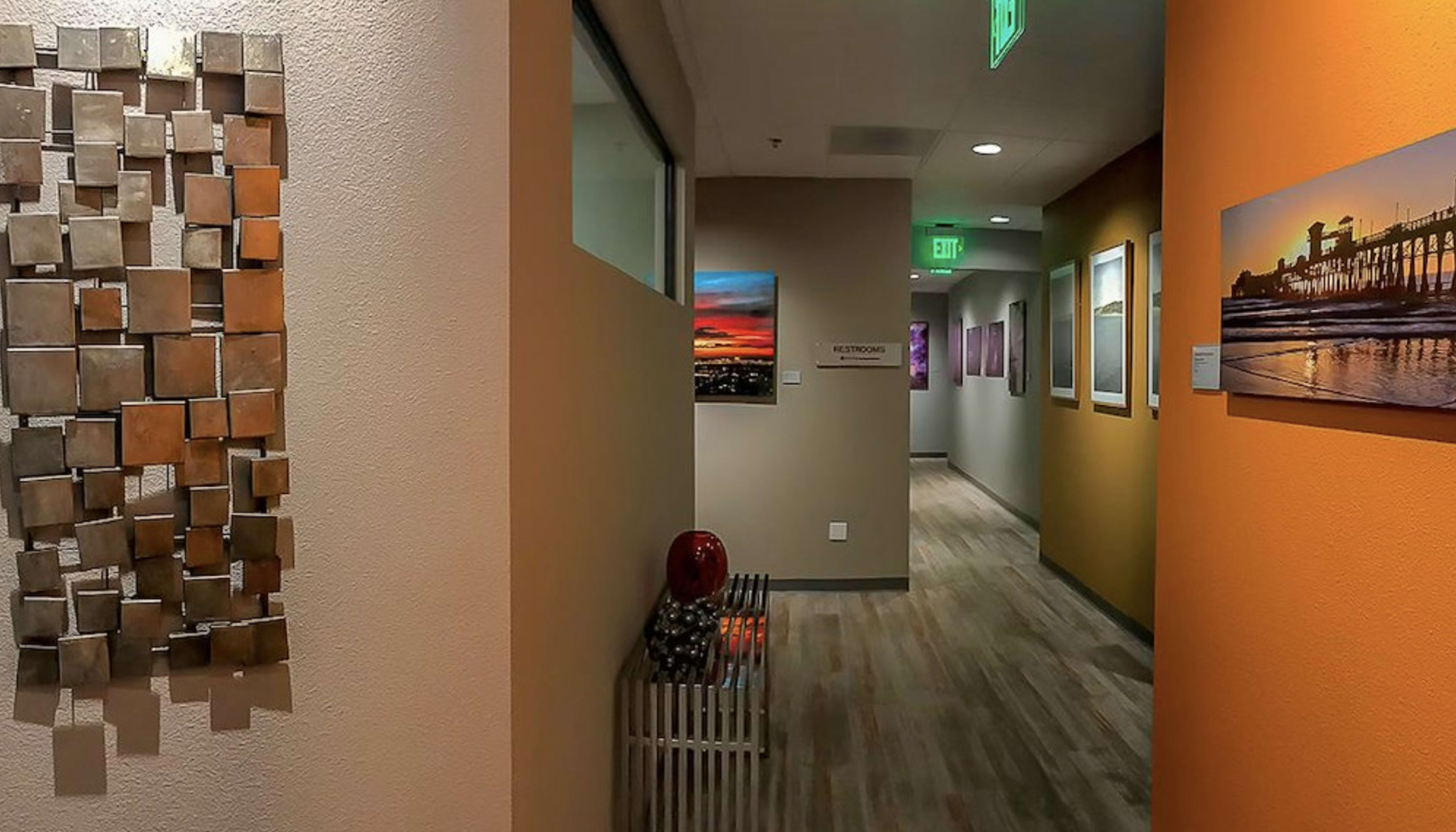 The image shows a well-lit hallway with framed artworks on the walls, a sign for restrooms, and a decorative metal wall piece on the left.