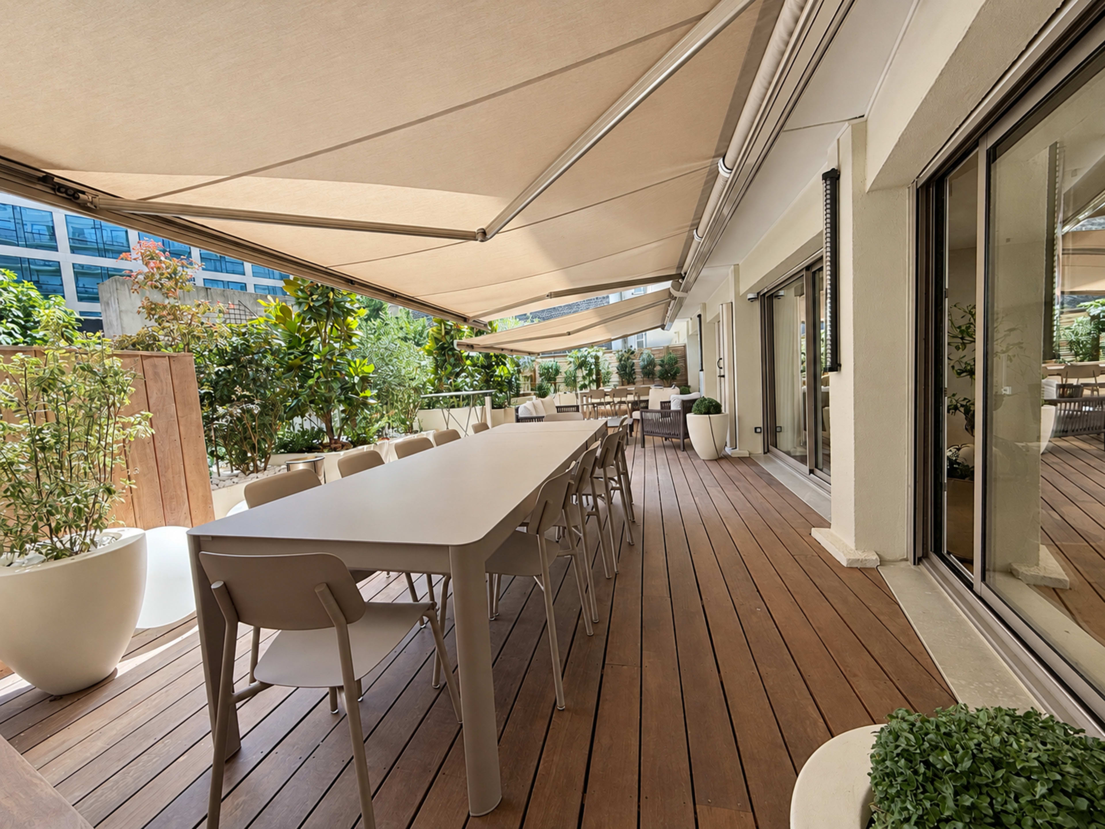 The image shows a modern outdoor dining area with a long table, several chairs, and large potted plants, shaded by a retractable awning.